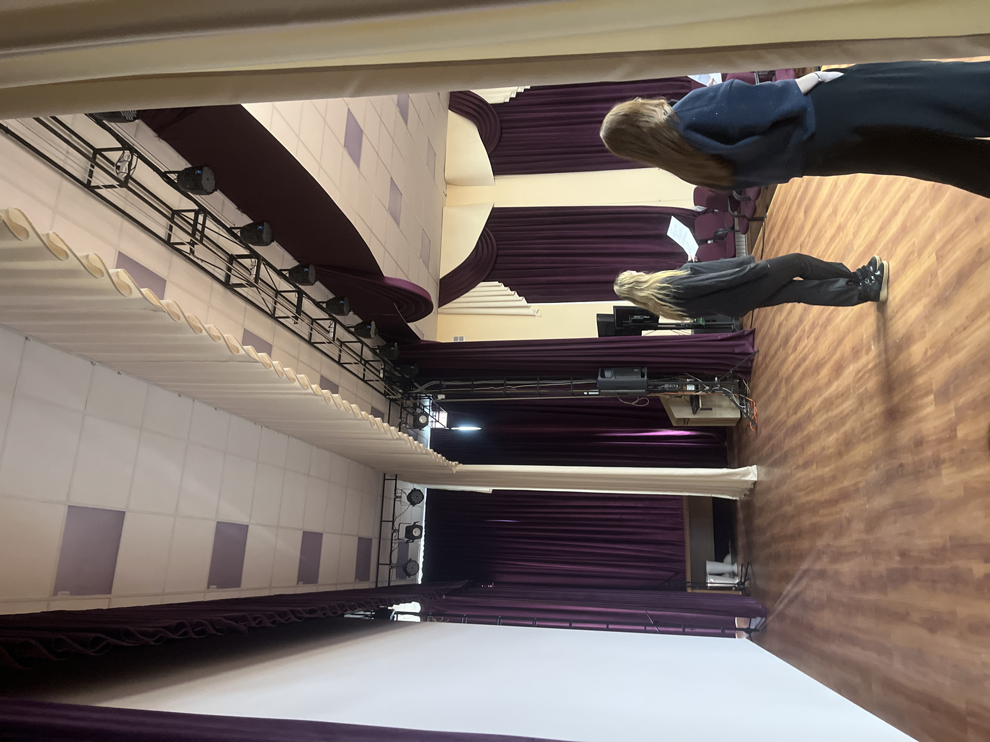 The image shows the interior of a theatre in an unknown location but presumed to be Saint Petersburg, Russia. In the foreground, the bottom edge of a large, white projection screen is visible, partially obscured by a purple curtain. Above the screen, a complex lighting rig is mounted on a black frame, with several adjustable spotlights. Behind the screen, purple stage curtains hang in vertical folds, partially concealing the backstage area.
In the midground, two individuals are standing. One has blonde hair and is wearing a grey hoodie and black pants and the other has red hair and is wearing a blue hoodie and black pants.
The floor is made of wooden planks. In the background, a few chairs can be seen. The walls are mostly white with purple accents.