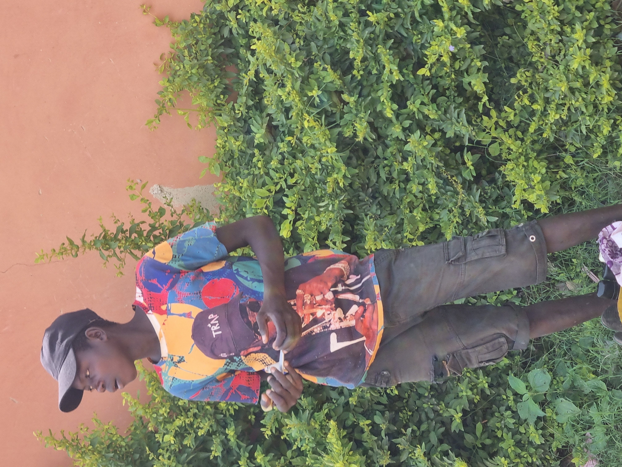 In Gwong, Nigeria, a young Black male stands outdoors, facing left and looking away from the camera. He is wearing a black baseball cap, a multi-colored t-shirt with graphic prints, and dark cargo shorts. His hands are positioned in front of him, holding what appears to be a cigarette and a lighter. Behind him, there is a dense green hedge and a peach-colored wall. The ground in front of the hedge is covered with grass and some debris. The lighting suggests it is daytime.