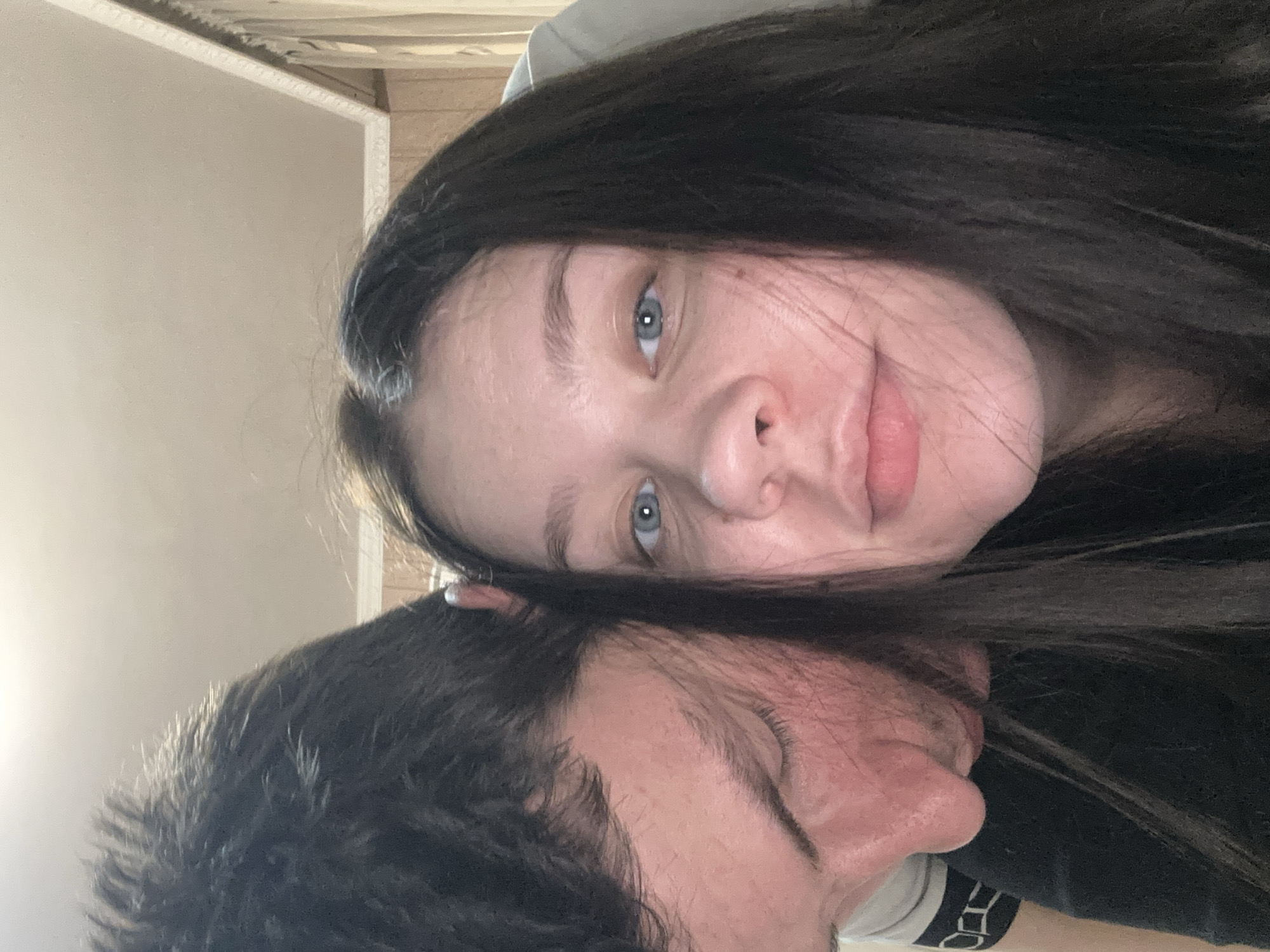 The image is a selfie featuring two people. A woman with long, dark hair and blue eyes occupies the top half of the frame. The lower half of the frame shows a man with dark hair and closed eyes. His face is partially obscured by the woman's hair. The background consists of a neutral-colored wall. There is no evidence to suggest the image was taken in Varna, Bulgaria.