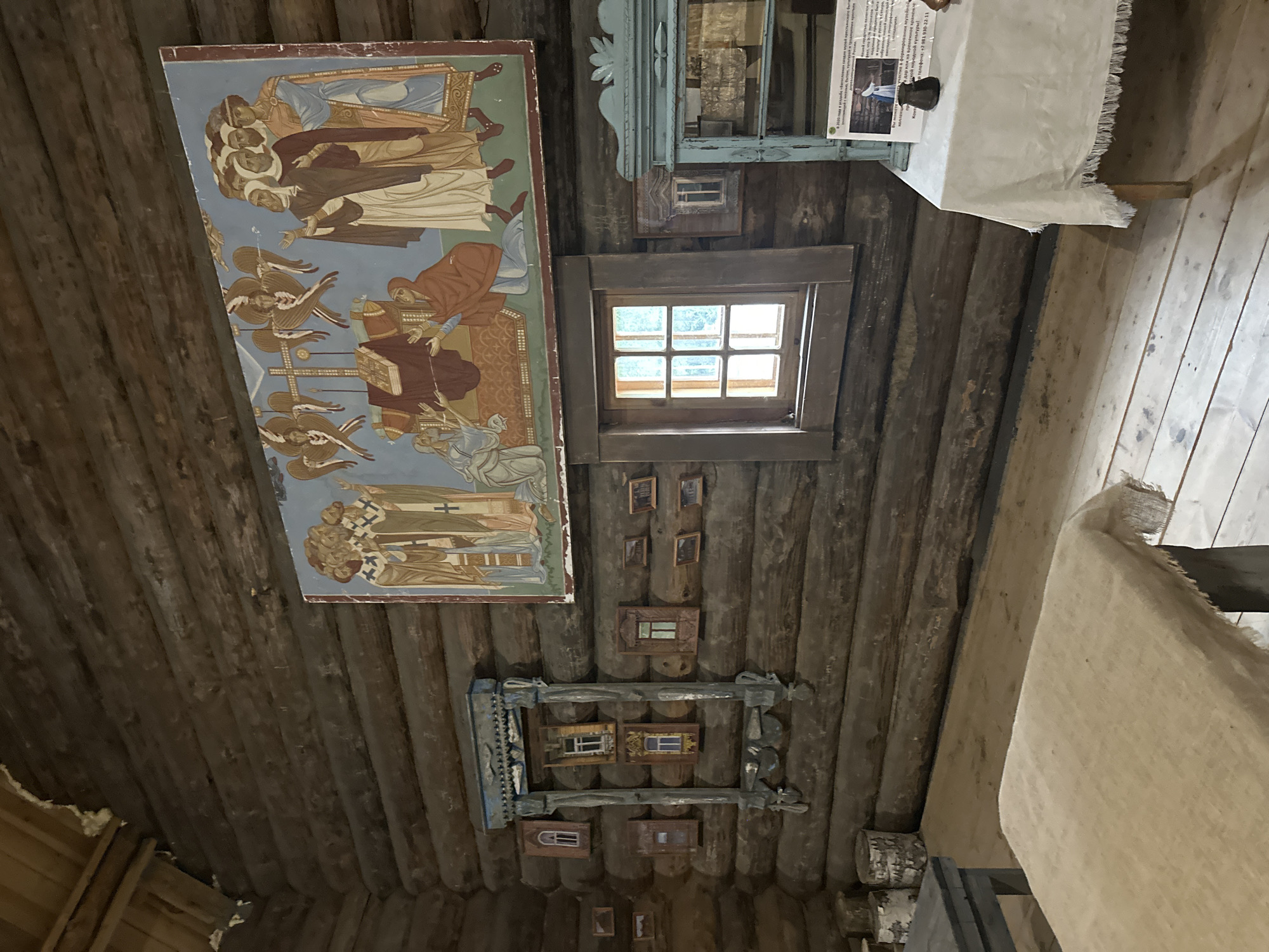 The image shows an interior view of a rustic wooden room, likely a museum exhibit in Nevskii Parkleskhoz, Russia. The walls are constructed from thick, horizontal logs.
Dominating the upper portion of the wall is a large, colorful mural depicting a religious scene. It shows several figures in traditional robes, some with halos, gathered around a central altar or table. Two winged figures, possibly angels, are also visible in the mural.
Below the mural, a decorative wooden frame with carved details is mounted on the wall. Within this frame, and also scattered around it, are several small, framed pictures of windows. These appear to be miniature decorative elements.
A wooden window with multiple panes is set into the log wall. To the right of the window is a light blue, ornate cabinet or shelving unit. A white tablecloth covers a table in front of the cabinet. A sign with text and an image is placed on the table, likely providing information about the exhibit.
In the foreground, a wooden table is covered with a rough, beige cloth. Another dark-colored table is visible to the left. Several short, cylindrical wooden logs, possibly firewood, are stacked on the floor in the background to the left. The floor is made of wooden planks. The overall impression is of a historical, culturally significant space.