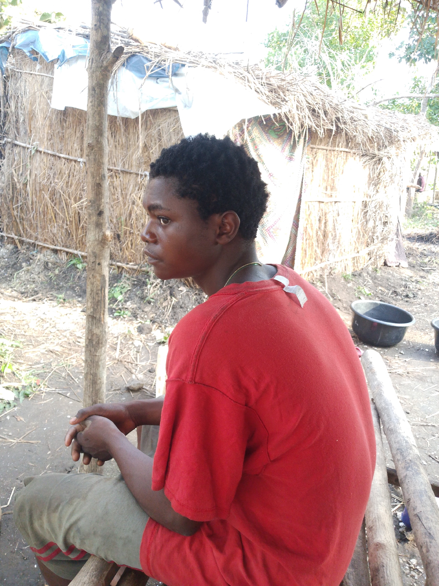 In the city of Kajola, Nigeria, a young person with dark skin and short black hair sits facing left, partially obscured by the edge of the frame. They are wearing a bright red t-shirt and light-colored pants with red stripes. Their hands are clasped in their lap. The background consists of a rustic hut made of thatch and branches, with some blue tarp visible on the roof. A wooden post stands to the left of the person. The ground is covered in dirt, dry leaves, and twigs. To the right, a black plastic basin filled with water rests on the ground. The scene appears to be outdoors, likely in a rural or semi-rural area.