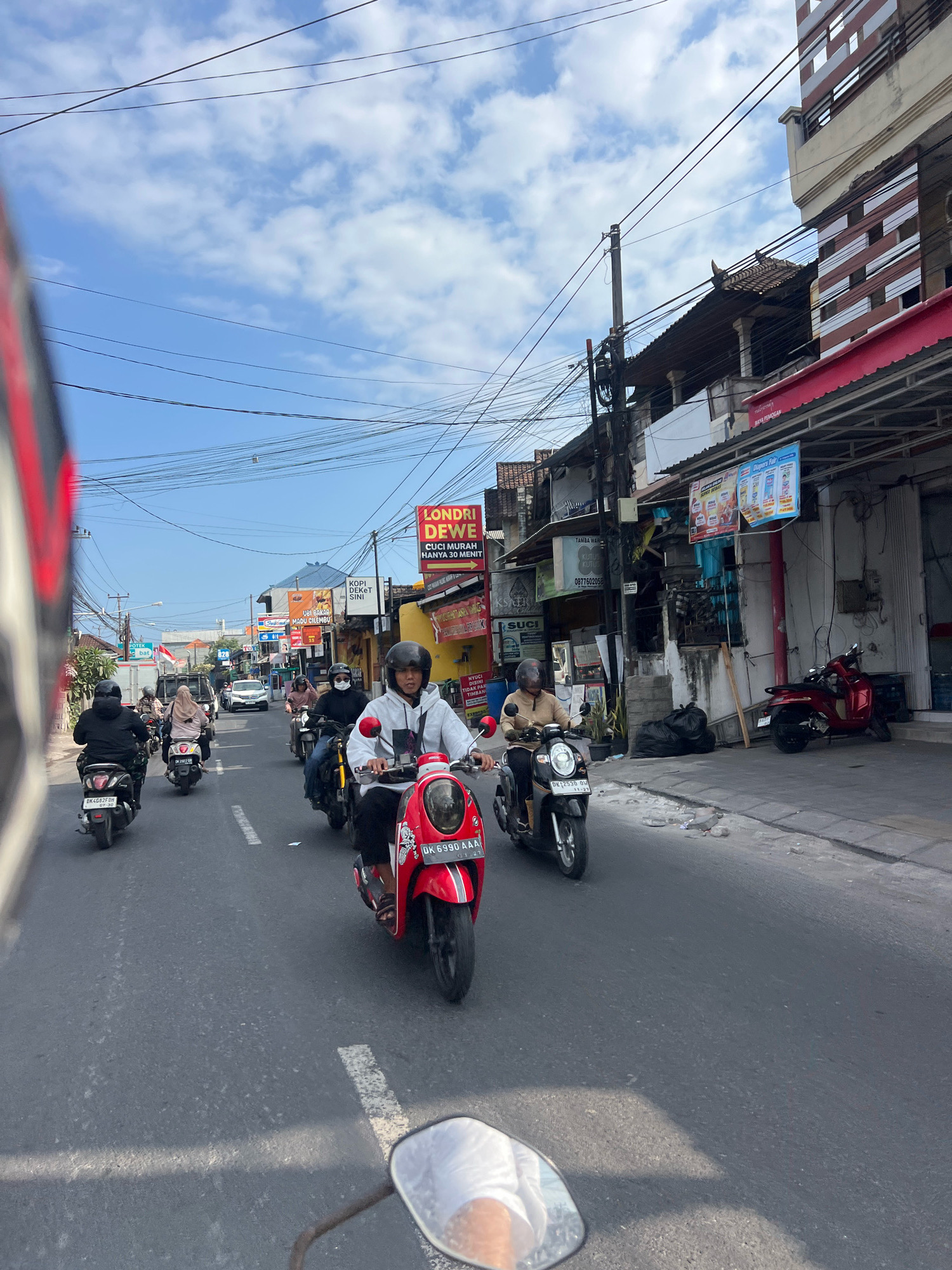 The image depicts a street scene in Denpasar, Indonesia, with multiple motorcycles navigating the road under a partly cloudy sky. The perspective suggests the photographer is also on a motorcycle, with a portion of their vehicle's handle and mirror visible in the foreground.
On the road, several individuals are riding motorcycles. In the immediate foreground on the right, a person in a white hoodie is riding a red scooter. Further ahead, to the left, another person in a light brown jacket is riding a black scooter. Behind them, more motorcycles and riders are visible, including one in a black outfit on a black scooter and another in a light brown hijab on a white scooter. A red motorcycle is parked on the sidewalk to the right.
The environment consists of a paved road with white lane markings. Along the sides of the road, there are buildings with shops and signs. Prominent signs include "LONDRI DEWE" with an offer for laundry services, and others with Indonesian text advertising various businesses. Power lines crisscross the sky above the street. The overall atmosphere suggests a busy, everyday scene in a commercial area of the city.