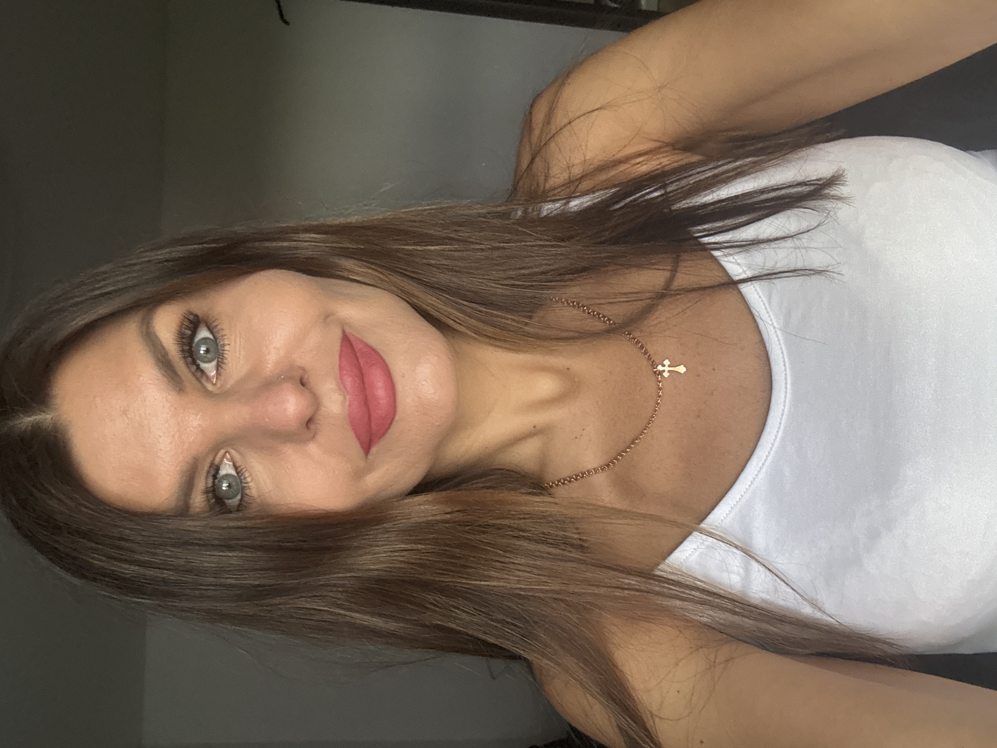 A woman with brown hair, gray eyes, and pink lipstick is wearing a white tank top and a gold cross necklace. She is indoors, possibly in Chaikovsky, Russia, although there is nothing in the image to definitively confirm that location. The background is a plain, light-colored wall.