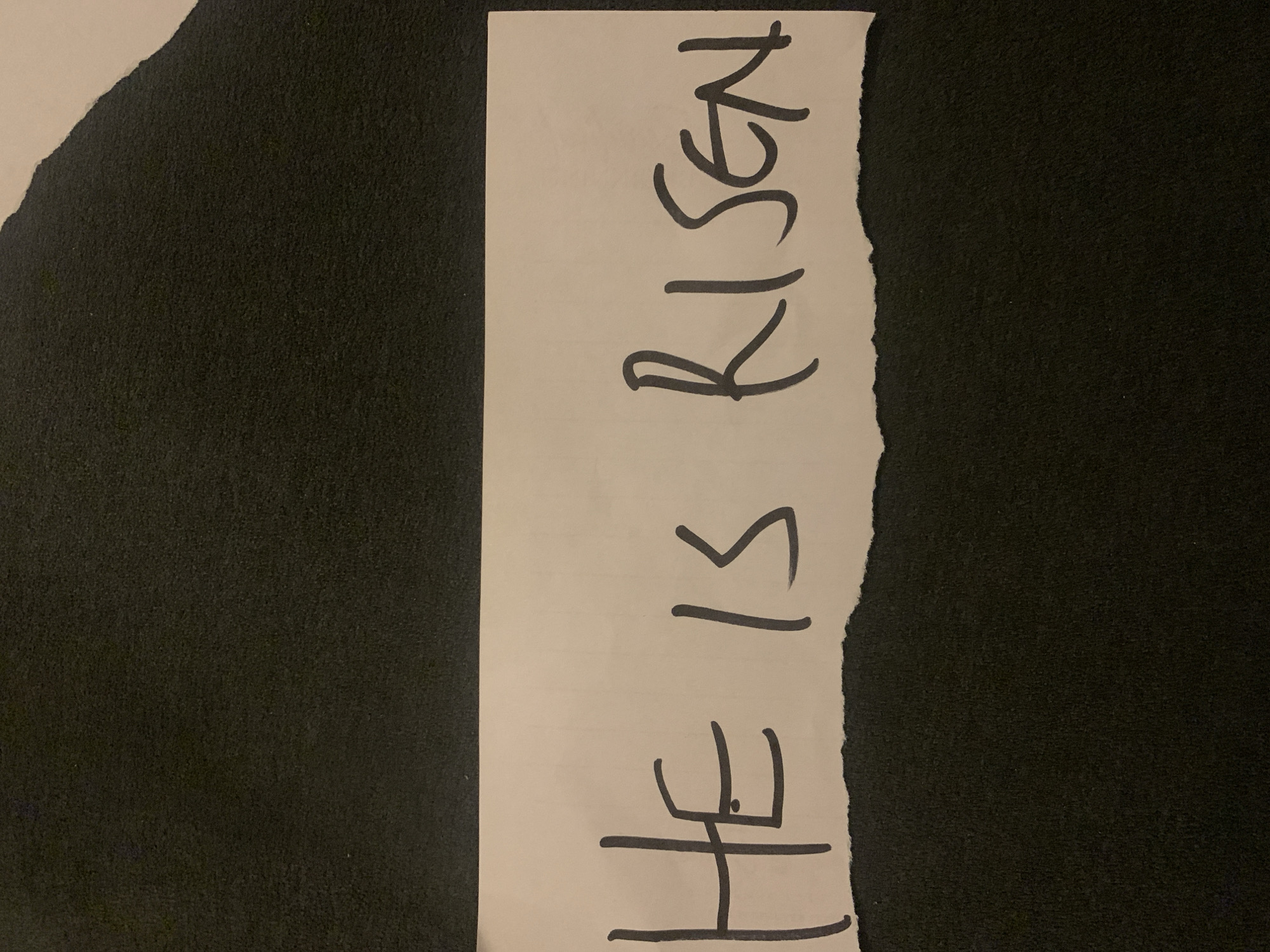 A piece of torn white paper with the handwritten message "HE IS RISEN" in black marker lies on a dark, textured surface, likely fabric. The message is vertically oriented. There are no people visible in the image. The location, Barakin Akawo, Nigeria, cannot be determined from the provided image.