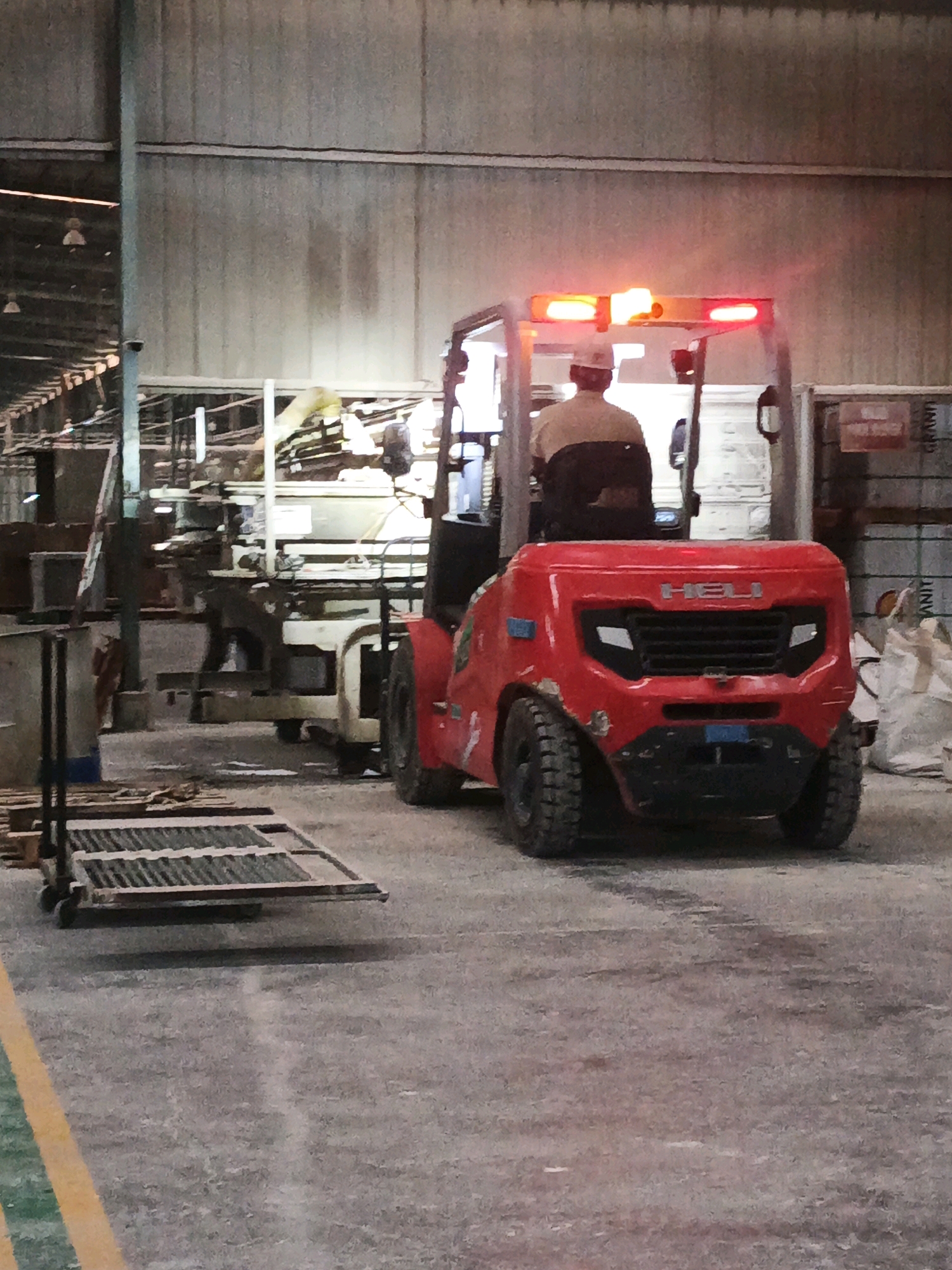 The image shows a red HELI forklift with a person in a white hard hat and a brown shirt sitting in the driver's seat, facing away from the camera. The forklift has flashing red lights on its overhead guard. In the background, there are industrial racks, various materials, and what appears to be a warehouse or factory environment. To the left of the forklift, a metal cart with wheels and a slatted top is positioned on the concrete floor. The scene suggests an active industrial setting, possibly within a logistics or manufacturing facility in Ngoro, Indonesia.