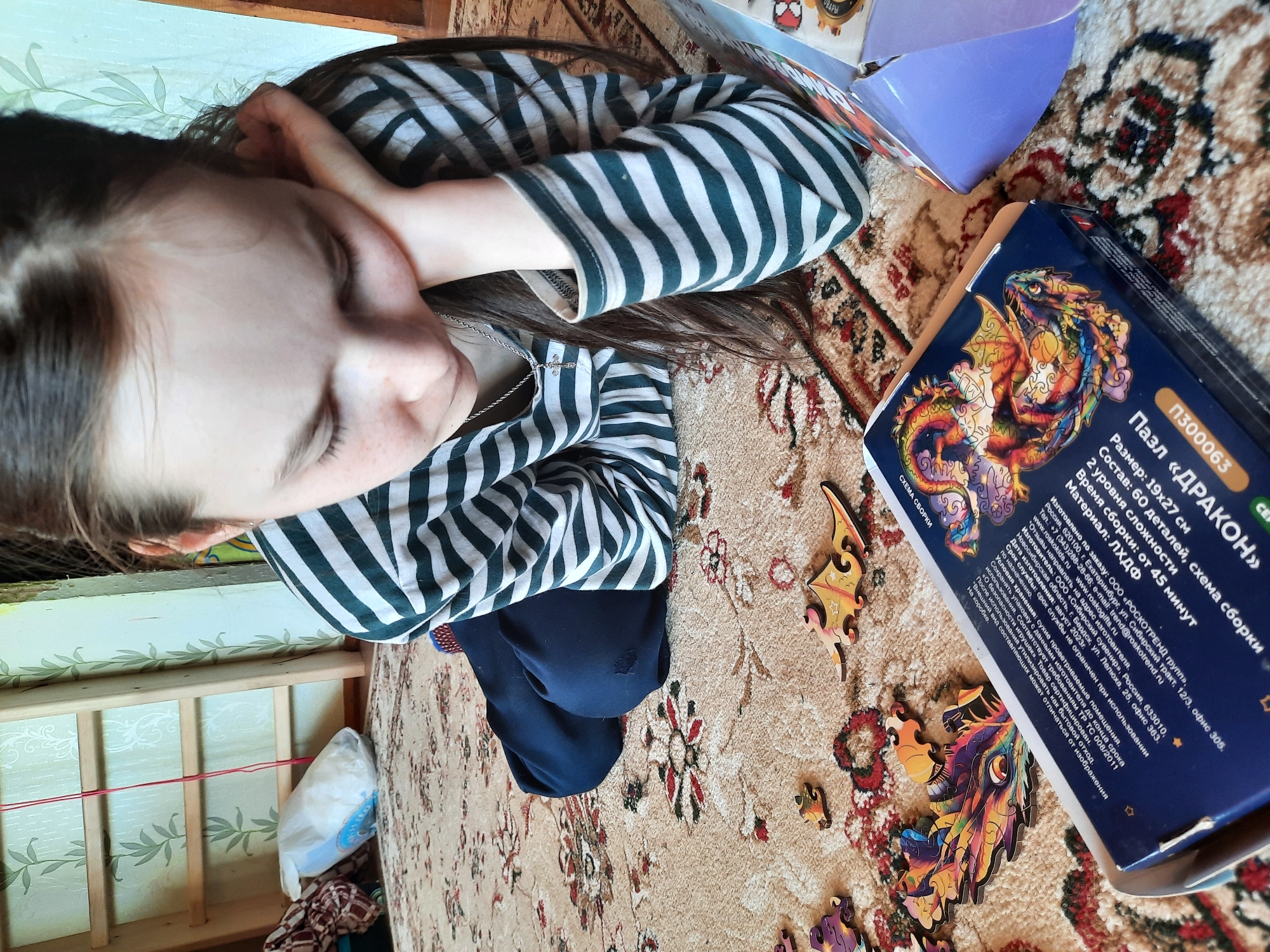 A young girl is lying on a rug, with her head resting on her hand. She is wearing a striped shirt and dark pants. Beside her is a puzzle box labeled "П300063 Пазл «ДРАКОН»" which translates to Puzzle "Dragon". Scattered around the box are several puzzle pieces. The rug is beige with floral and geometric patterns in red and brown. In the background, there is a wooden structure with a decorative pattern and a plastic bag containing what appears to be stuffing. The puzzle box indicates it was made in Berdsk, Russia, in August 2023. The image does not show any location in the city of Архангельское, Russia.