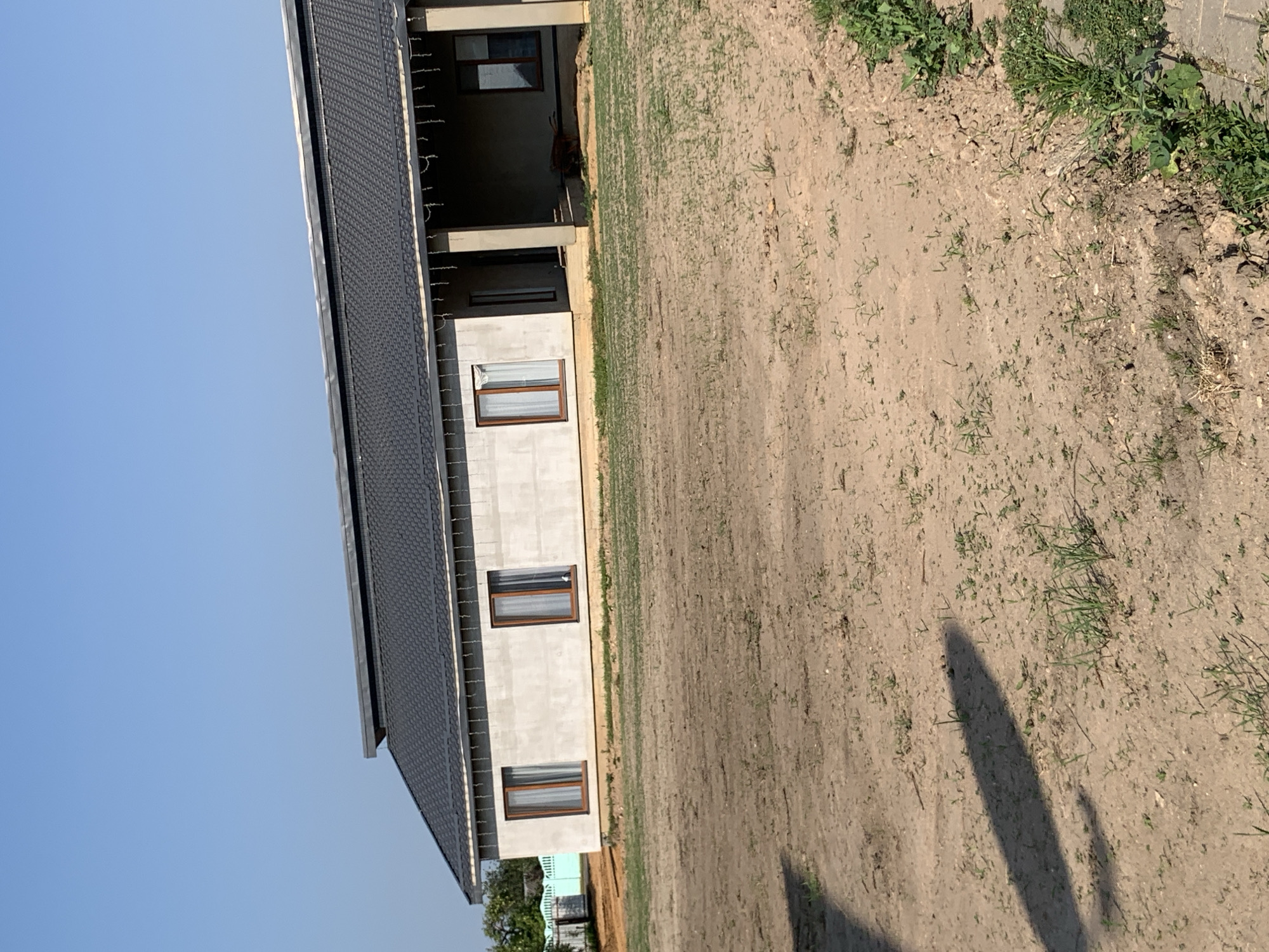 The image shows a residential building under construction in Kapyĺ, Belarus. The building is a single-story structure with a dark gray tiled roof. The exterior walls appear to be made of concrete blocks, with several windows that have wooden frames and sheer white curtains. A covered porch extends along a section of the building. The ground in front of the house is uneven and covered in dirt and sparse vegetation. A shadow of an object, possibly a tree or another structure, is cast across the foreground. In the background, more construction materials and fencing are visible, along with some trees. The sky is clear and blue. The scene depicts a rural or suburban area during the daytime.