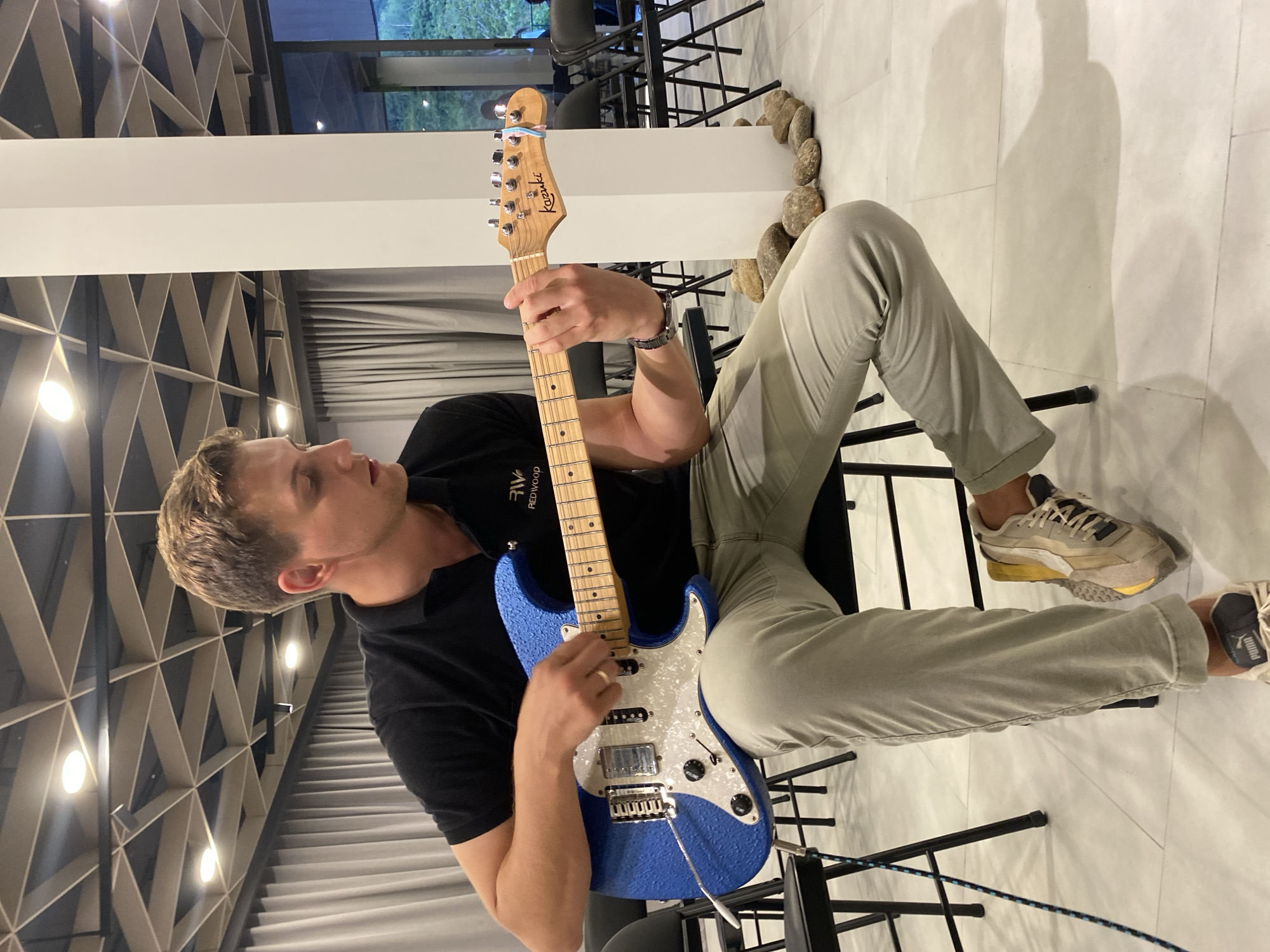 A man is depicted sitting and playing a blue electric guitar in what appears to be a modern indoor space. He is focused on the instrument, looking down with a slightly open mouth, indicating concentration or singing. He wears a black short-sleeved top with "Redwood" partially visible on the collar, light grey or khaki pants, and light-colored sneakers with "PUMA" visible on one. A watch is on his left wrist.
The electric guitar is a vibrant blue with a textured finish, a white pearloid pickguard, chrome hardware including a tremolo bridge, and a light wooden neck and headstock. The brand name "Kazuki" is clearly visible on the headstock.
The background features a white tiled floor and a prominent white pillar. A decorative arrangement of smooth, natural stones lines the base of a wall or pillar. Above, a striking geometric pattern of light-colored wood or material covers the ceiling or upper wall, with circular light fixtures embedded within. Through large windows in the distance, dark foliage and greenery are visible, suggesting an outdoor natural setting. The ambient lighting suggests it is evening or night. The scene is set in Choeng Thale, Thailand.