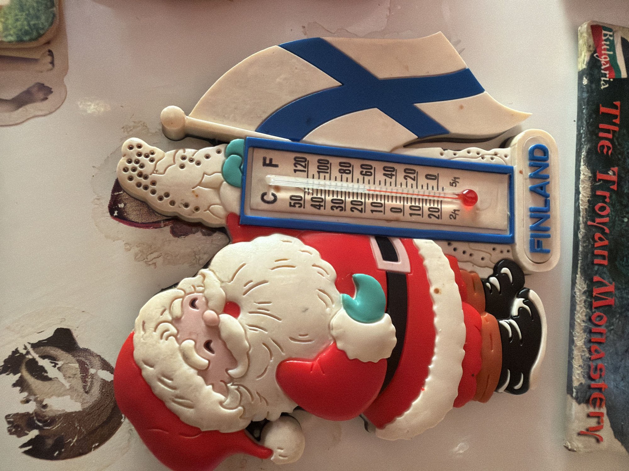 A close-up, slightly angled, studio shot displays a Christmas-themed refrigerator magnet. The magnet features Santa Claus alongside the Finnish flag and a thermometer reading around 50 degrees Fahrenheit. Adjacent to the magnet is a book titled "The Troyan Monastery, Bulgaria." The scene lacks contextual details such as time, location, and weather conditions. No interactions or subjects besides the inanimate objects are present.