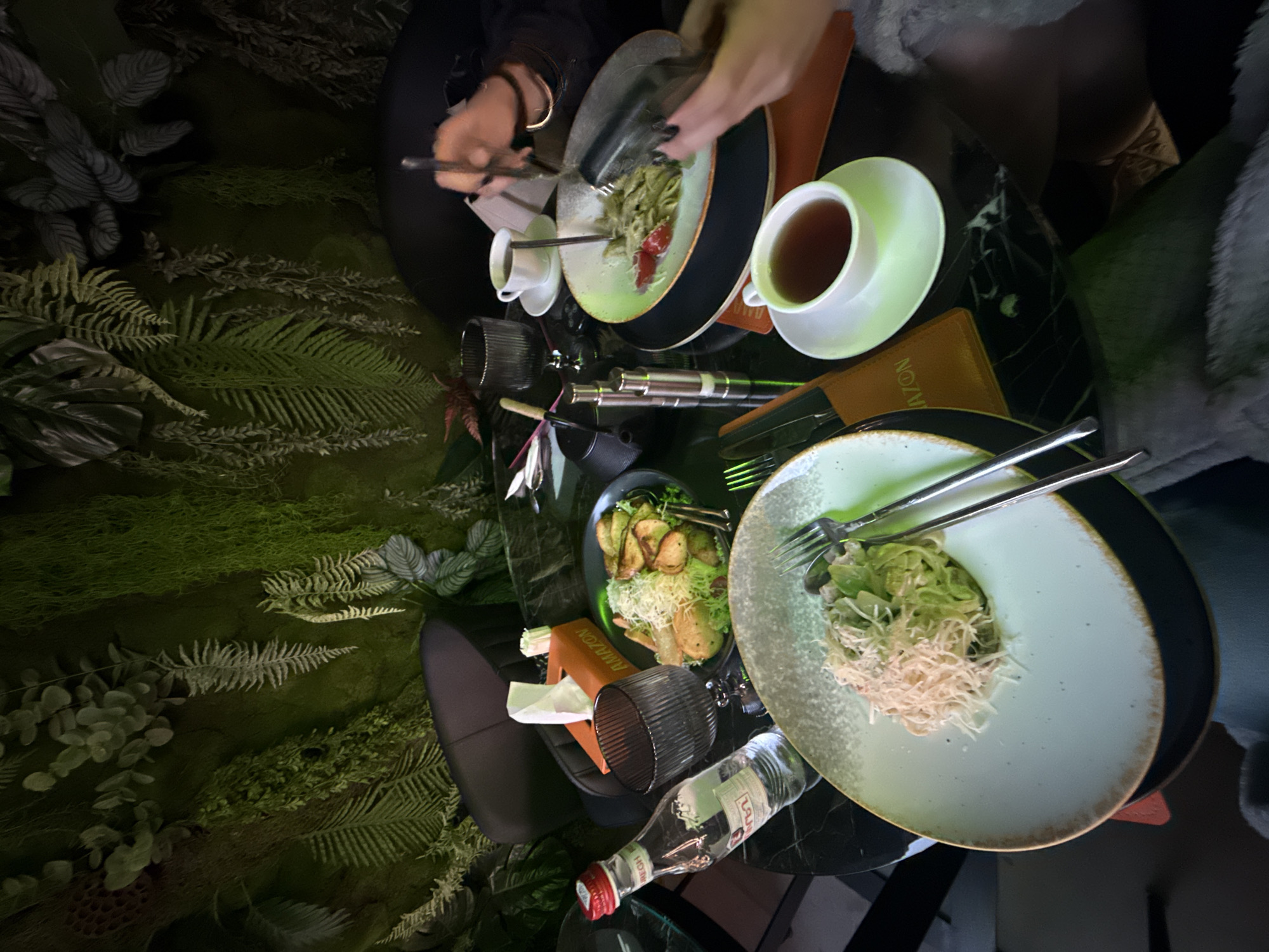 A top-down view reveals a meal at NOZOMI Yerevan, Armenia. Two people are seated at a black, marbled table with plates of food. One plate holds green pasta with red sauce, being eaten by a person using utensils. A cup of tea sits next to the plate. The other plate contains greens and fried pieces of food, with a water bottle and tissue box nearby. Lush green plants form the backdrop on the left.