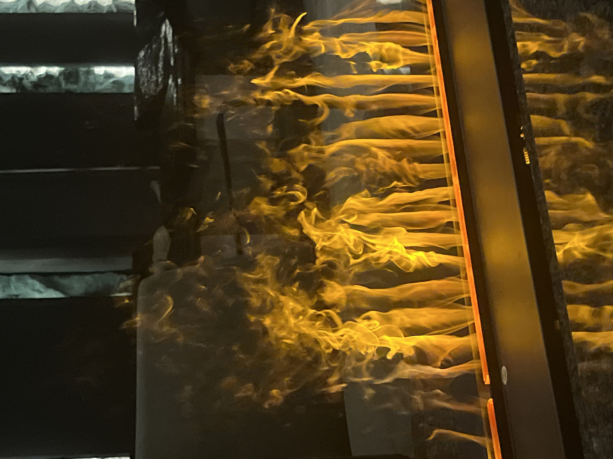 The image displays a modern, linear fireplace featuring vibrant orange and yellow simulated flames rising vertically from a dark, horizontal unit. A narrow, illuminated orange strip is visible at the base of the flames. The artificial fire is reflected on a dark, polished surface directly beneath the unit. The background is dimly lit and out of focus, revealing blurred vertical architectural elements on the right, suggestive of textured panels or columns. No individuals or other discernible subjects are present within the frame. The scene is an interior space in Sofia, Bulgaria.