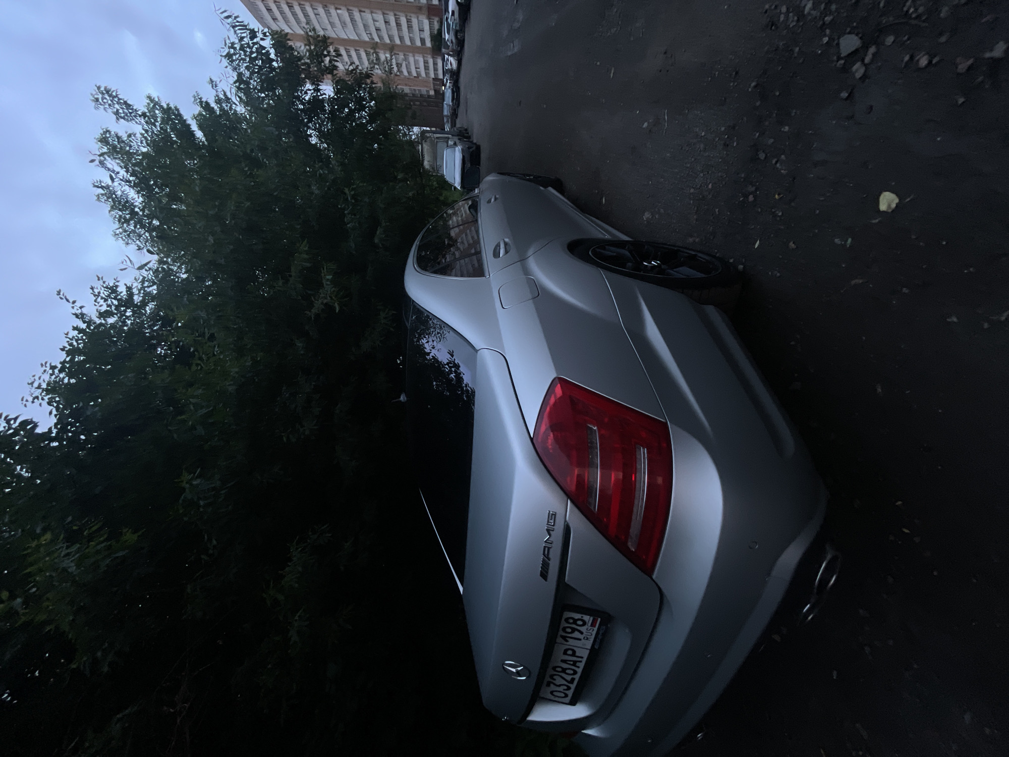A silver Mercedes-Benz S-Class sedan is parked on a dark, wet asphalt surface next to a dense line of trees. The car's rear is visible, featuring a license plate that reads "0328AP 198 RUS" and an "AMG" emblem. The vehicle has tinted windows and black rims. In the background, apartment buildings and other parked cars are discernible, indicating an urban environment. The sky is overcast with a cloudy, grayish hue. The lighting suggests it is either early morning or late evening.