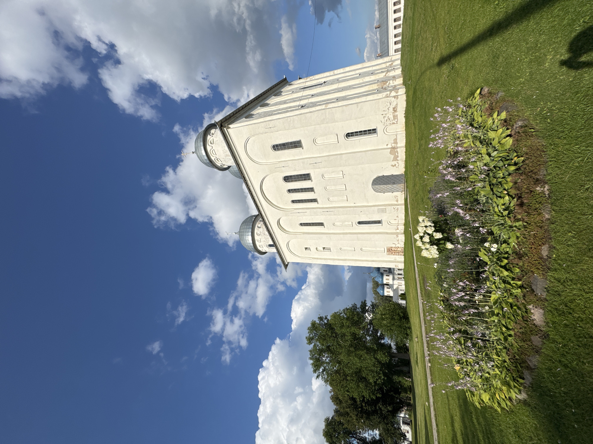 The image depicts a bright, sunny day with a blue sky filled with fluffy white clouds. In the foreground, a bed of green plants with delicate purple flowers is in bloom. Behind the flowerbed, a large, white, stone building with a grey domed roof stands on a grassy hill. The building has several arched windows and a large, metal-clad door. In the background, more white buildings with blue domed roofs can be seen. The scene is set in the city of Veliky Novgorod, Russia, likely within the grounds of the Saint Sophia Cathedral or another significant historical site, given the architectural style and the presence of multiple domed structures. There are no people clearly visible, but a shadow of a person is cast on the grass to the right of the frame. The grass is well-maintained and slopes down from the building.