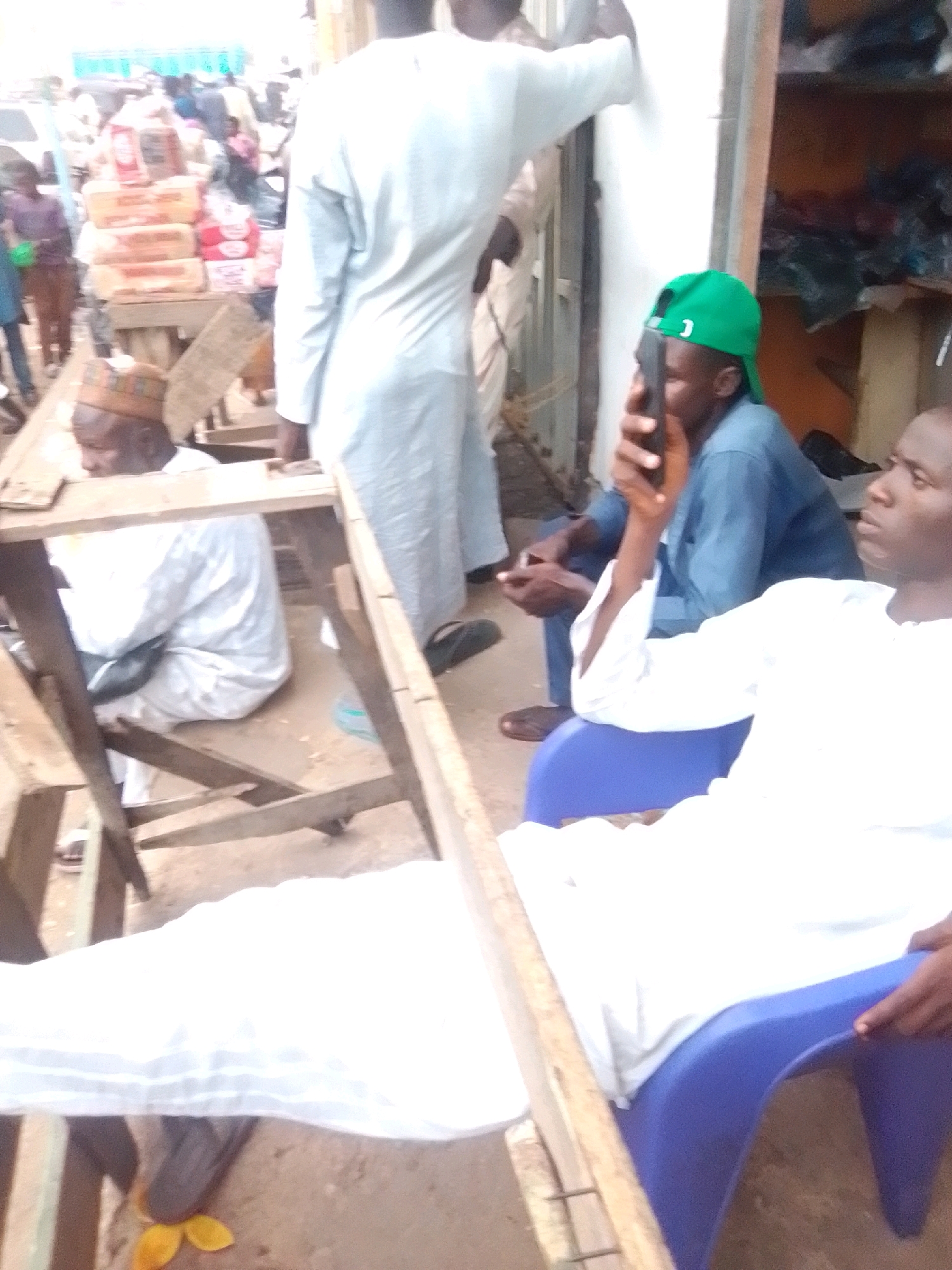 The image shows an outdoor market scene in Arawa, Nigeria. Several individuals are present, some of whom are wearing traditional Nigerian attire. In the foreground, a person in a white robe is seated on a blue chair, partially obscured by a wooden structure. To the left, another individual in a white robe is seated. In the middle ground, a person wearing a green cap and a blue shirt is holding a mobile phone to their ear. Behind them, a stall is visible with stacks of goods, including what appear to be packaged food items. The overall environment suggests a bustling marketplace with various goods and people.