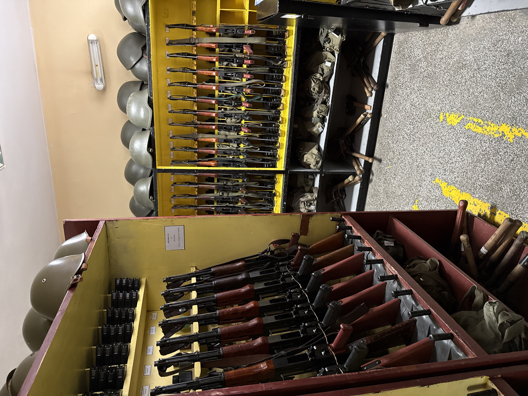 The image shows a room in an unknown location containing a storage unit with several shelves. The unit is filled with military equipment, including a row of helmets, several rows of rifles, and bags. The floor is concrete with yellow painted markings.