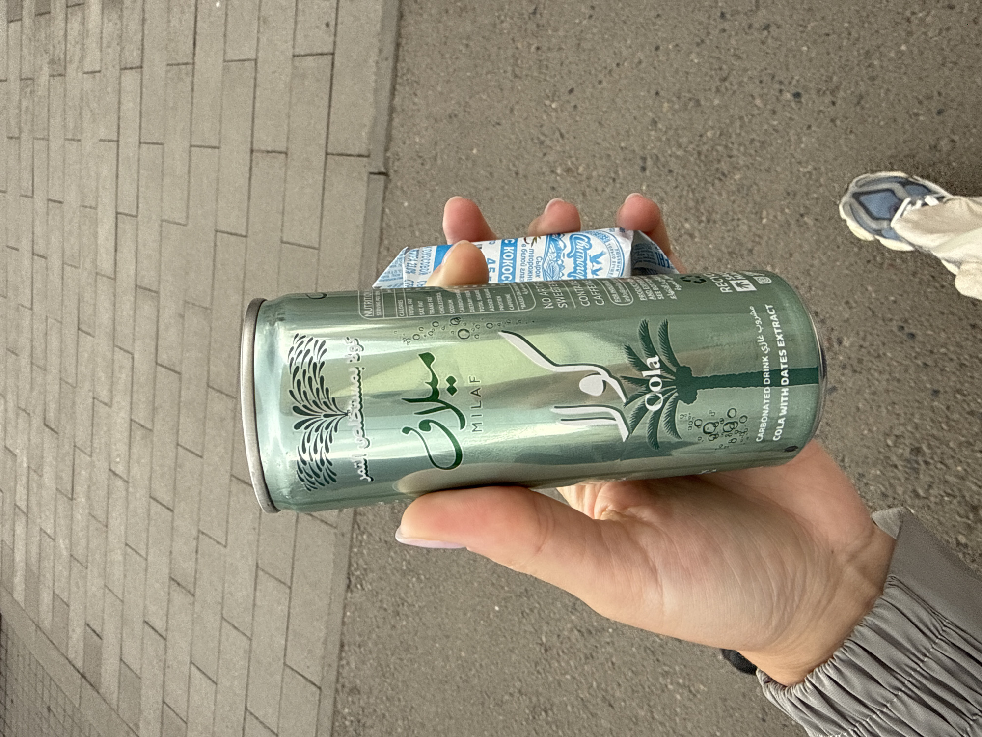 A hand holds a can of "Milaf Cola with Dates Extract," along with a "Koloskovsky c Kokos" curd cheese bar. The setting appears to be outdoors, possibly a street or sidewalk, as indicated by the pavement and a person's shoe in the background. The individual is wearing a gray sleeve. The brand of the cola is "Milaf" and the label also contains Arabic text. The curd cheese bar has Cyrillic text on it and the brand name "Koloskovsky". The image is taken at eye level with the subjects in the palm of the hand.