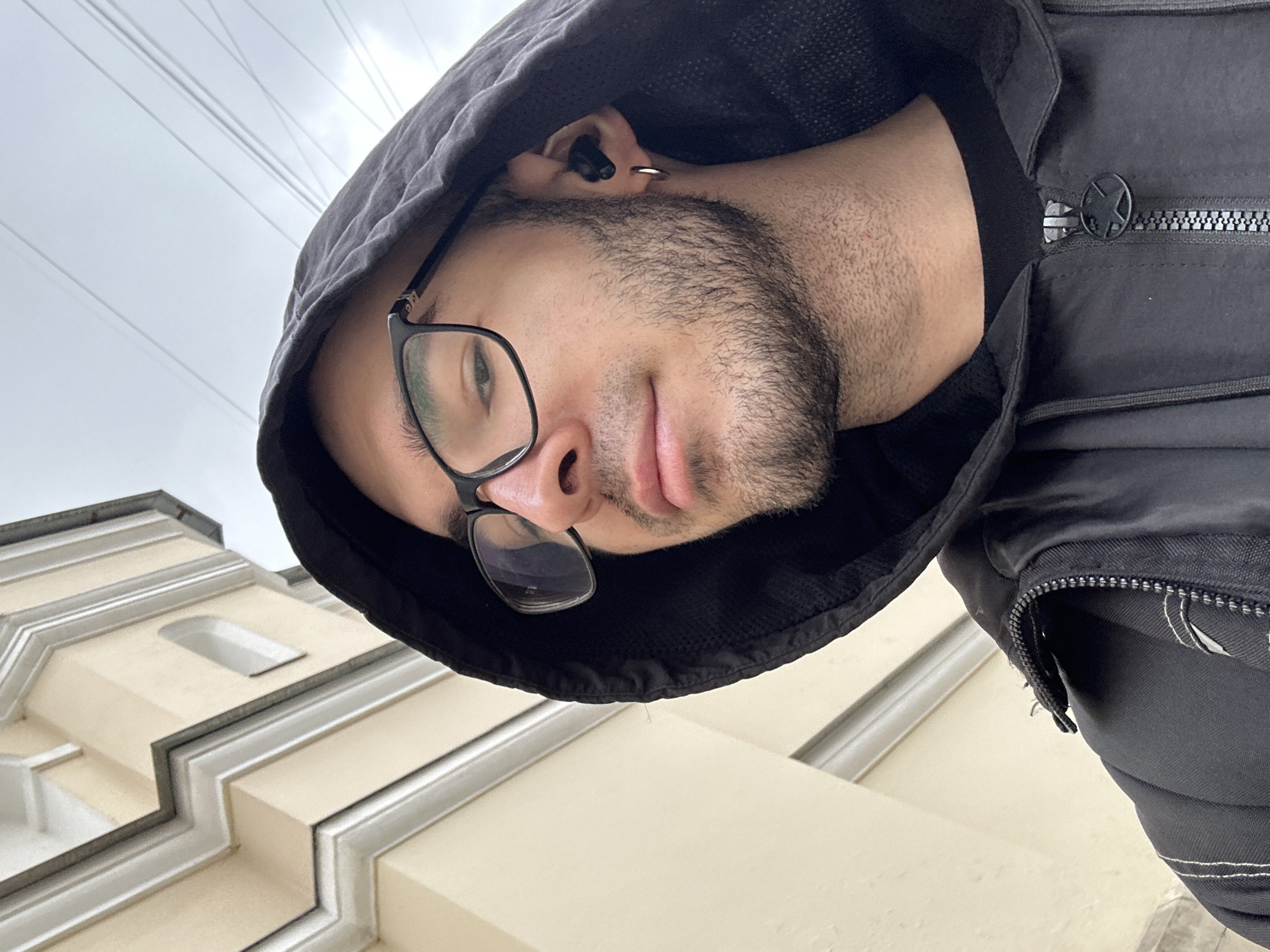 A medium shot taken from a low angle shows a man wearing glasses and a black hooded jacket. The man has a beard and an earring. Behind the man is a light-colored building with decorative white trim. Based on architectural characteristics, it's plausible this photo was taken in Moscow, Russia. Overhead wires are visible in the sky.