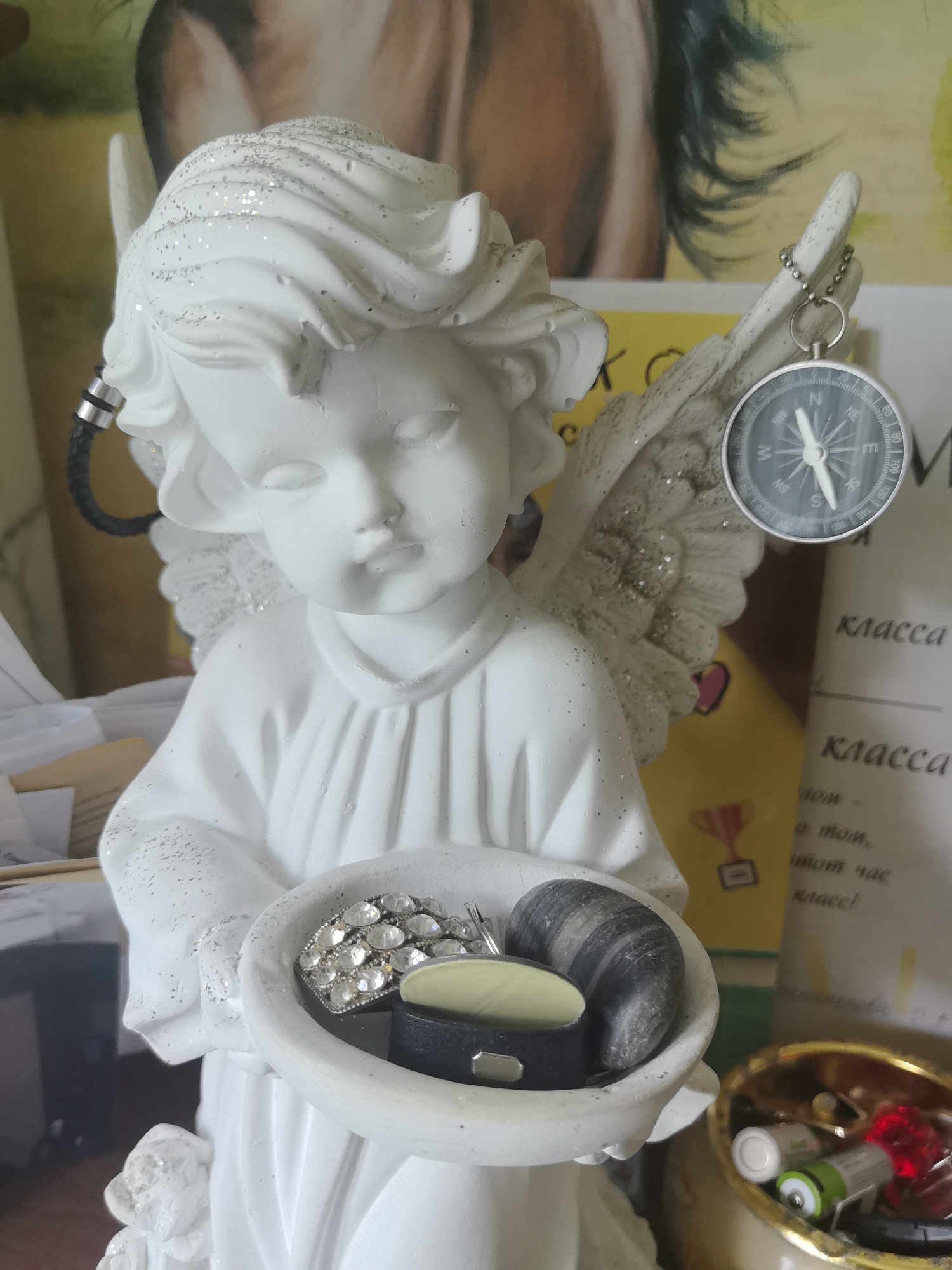 This is an indoor, eye-level, medium shot of a white, glitter-covered cherub figurine holding a bowl. The cherub's wings are also glittered, and it has a small compass hanging from a chain attached to its right wing. The bowl contains a sparkly ring, a smooth dark gray stone, and a black, rectangular object with a silver clasp. Behind the figurine, a blurred background shows a painting with a human figure, yellow and white papers with text, and a gold-colored bowl filled with batteries and a red object. The scene is lit from the front, casting a slight shadow on the cherub's face. The location is an indoor setting, possibly a home or an office, and the specific location in Homyel, Belarus cannot be determined from the visual information provided.