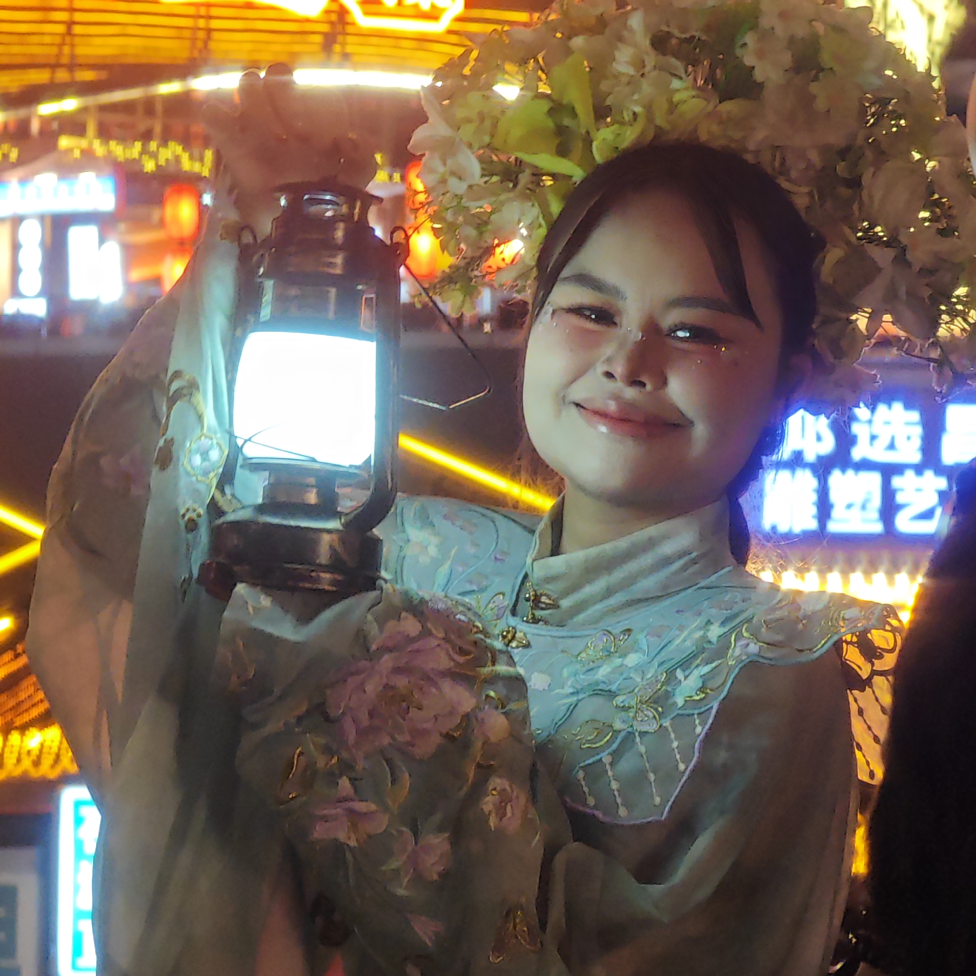 A person is holding a lantern in the Yuzhong District of China. The person is wearing an ornate garment and has flowers in their hair. The background is brightly lit with neon signs.