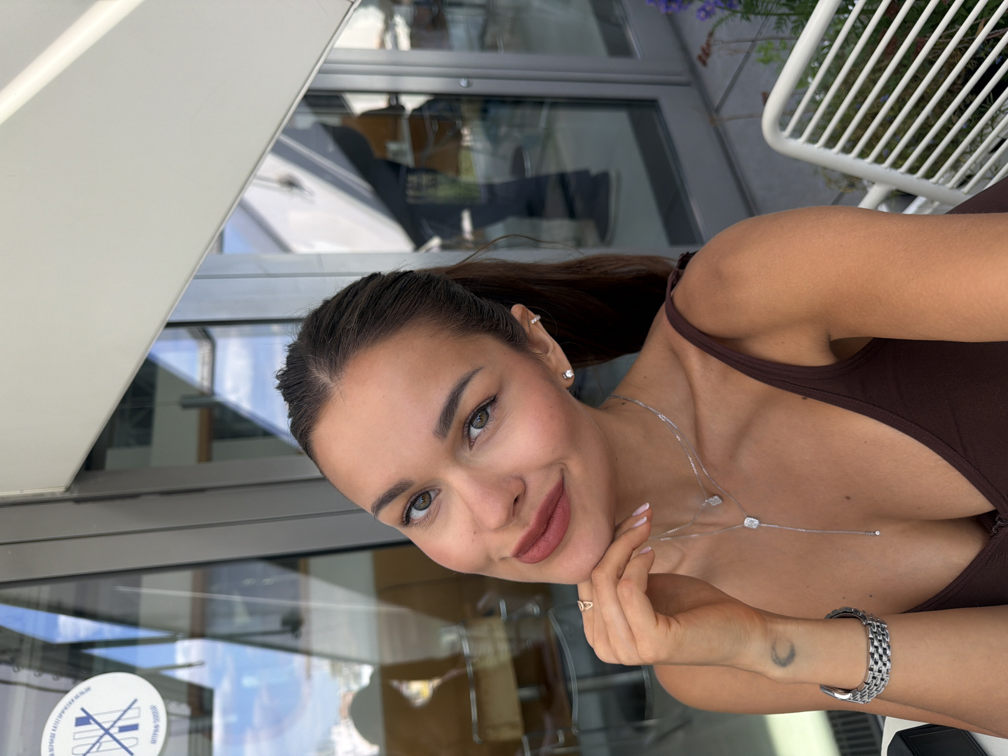A young woman with a gentle smile is the central subject of this close-up portrait, taken at an outdoor cafe or restaurant, likely in Moscow, Russia. She has medium-brown hair pulled back into a sleek ponytail, showcasing small diamond stud earrings. Her light brown eyes meet the camera, complemented by natural-looking makeup and a matte pinkish-brown lipstick. She wears a dark brown tank top or camisole and accessorizes with a delicate silver necklace featuring multiple square-shaped pendants that drape down her chest. A gold-toned ring adorns her right index finger, and a silver-toned watch with a link band is visible on her left wrist, above a faint crescent moon tattoo.
Her right hand is gently placed under her chin, adding to a relaxed and engaging pose. The background features large glass windows reflecting blue sky, clouds, and architectural elements, suggesting a bright, sunny daytime setting. Part of a white slatted outdoor chair and green foliage are visible to the right. On one of the glass panels, a circular sticker is affixed, displaying a symbol of crossed-out cutlery and a glass, indicating a 'no food or drink' policy, with text in Cyrillic script encircling the symbol.