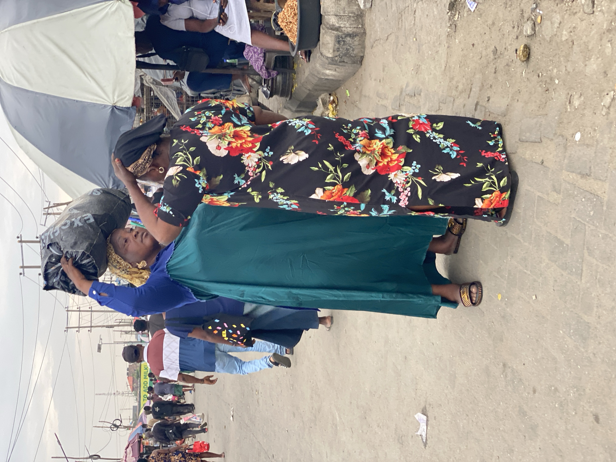 The image shows a busy street scene, likely a market, in Lekki, Nigeria. Two women are the primary subjects. One woman wears a dark dress with a bright floral pattern. The second woman is carrying a large, dark sack on her head and is wearing a blue blouse and a green skirt. Both women appear to be adjusting their head coverings. The street is crowded with people, and there are market stalls and umbrellas visible. The ground is a light grey pavement. Utility poles and overhead wires are present, and the sky is overcast.