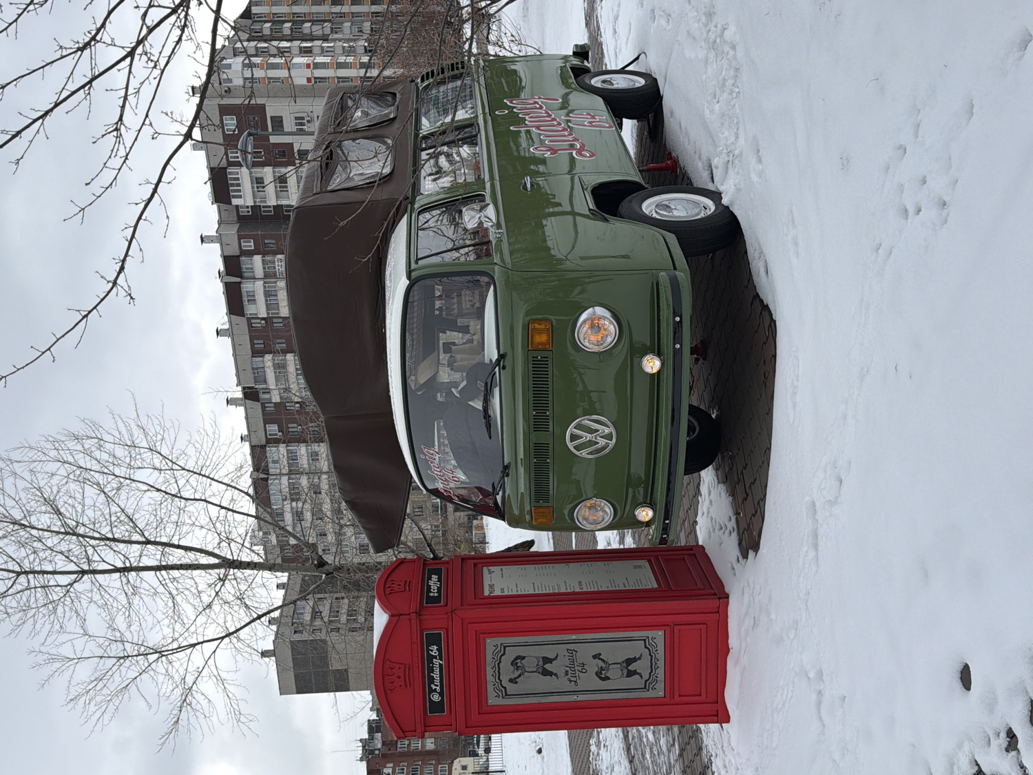 A vintage olive green Volkswagen Type 2 van, appearing to be a mobile coffee shop, is parked on a paved surface in a snowy, urban setting. The van features a white roof section and a brown canvas pop-top or awning extended over its driver's side. The words "Ludwig Coffee" are painted in red, stylized script on the side of the vehicle. The iconic Volkswagen emblem is prominently displayed on its front grille, and its headlights are illuminated.
Positioned to the left of the van is a distinctive red structure, resembling a British telephone booth, which serves as a menu or information stand. This stand also displays the "Ludwig Coffee" logo, featuring a pair of dancing or skating figures, alongside the text "#coffee" and "@Ludwig.64".
The ground to the right of the van and surrounding the red stand is covered in fresh snow, indicating a winter scene. Bare tree branches are visible against an overcast, grey sky. In the background, several large, multi-story apartment buildings line the horizon. The setting is a cold, daytime environment in an urban area of Krasnoyarsk, Russia.