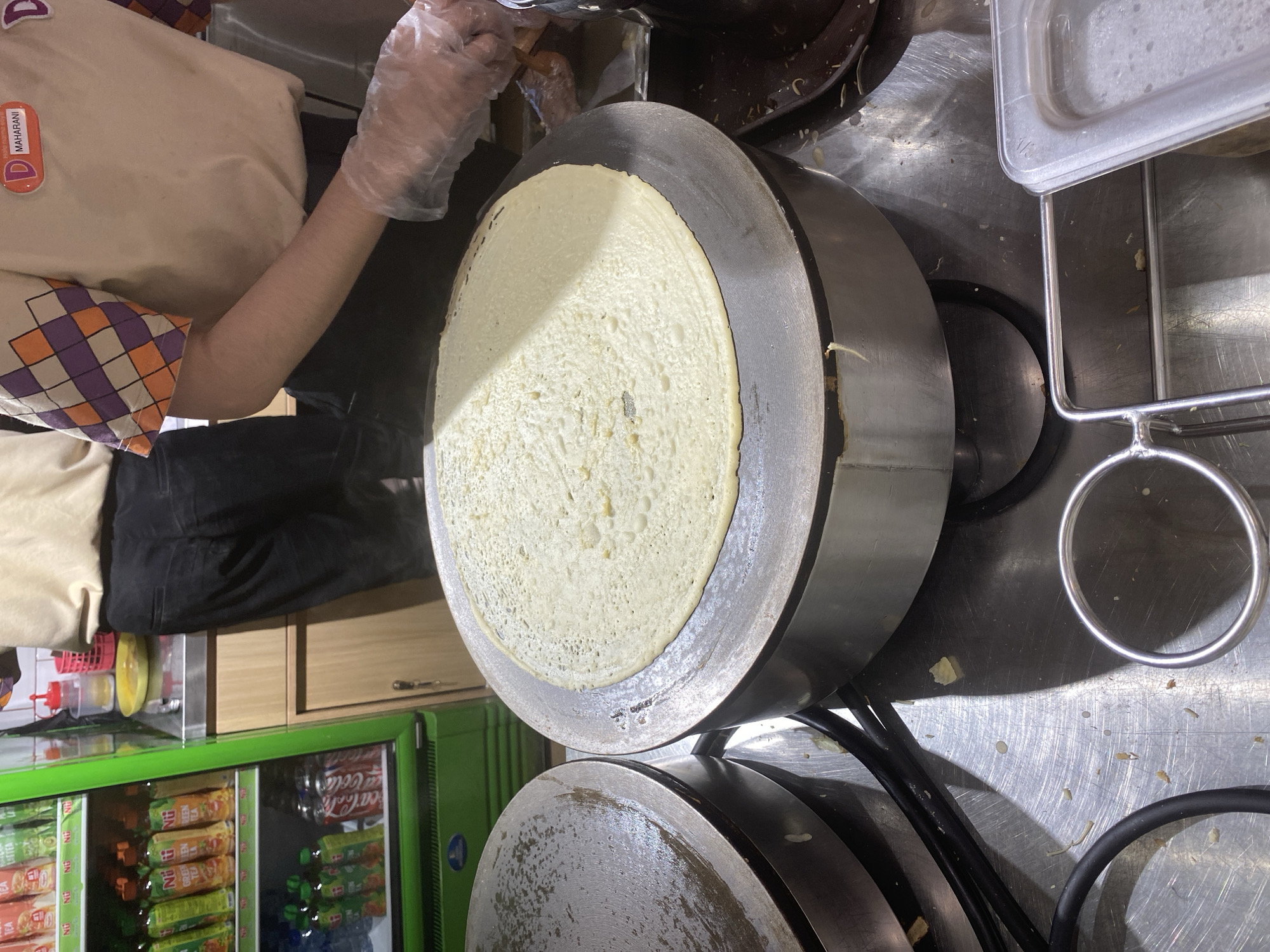 The image shows a close-up view of a crepe being cooked on a round, flat electric griddle at what appears to be a food stall. A person, wearing black pants and a tan shirt with a colorful checkered pattern on the sleeves and a name tag that reads "D MAHARANI," is visible from the waist down, their right hand, covered by a transparent plastic glove, is reaching towards the crepe. To the left of the griddle, another similar griddle is visible, appearing to be clean and possibly preheating. In the background, a bright green refrigerator is stocked with various bottled drinks, including "NÜ GREEN TEA" and Coca-Cola. The overall environment suggests a commercial food preparation setting, likely in West Jakarta, Indonesia, given the mention of the Maharani name tag, which is a popular chain in the region. The surfaces are metallic and reflective, indicating a professional kitchen or food service area.