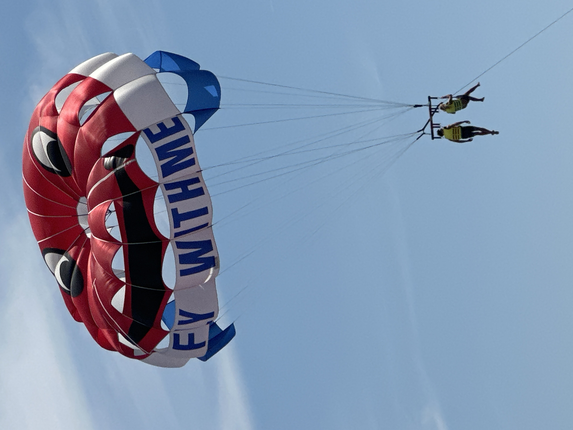 This is a low-angle, medium shot of two people parasailing over the city of Budva, Montenegro. The parasail is red, white, and blue, with a cartoon face design and the words "FLY WITH ME" printed on it in blue. The two individuals are wearing yellow life vests and are suspended from the parasail by ropes. The sky is clear and blue with a few wispy clouds. The scene captures the thrill and adventure of parasailing in a beautiful coastal setting.