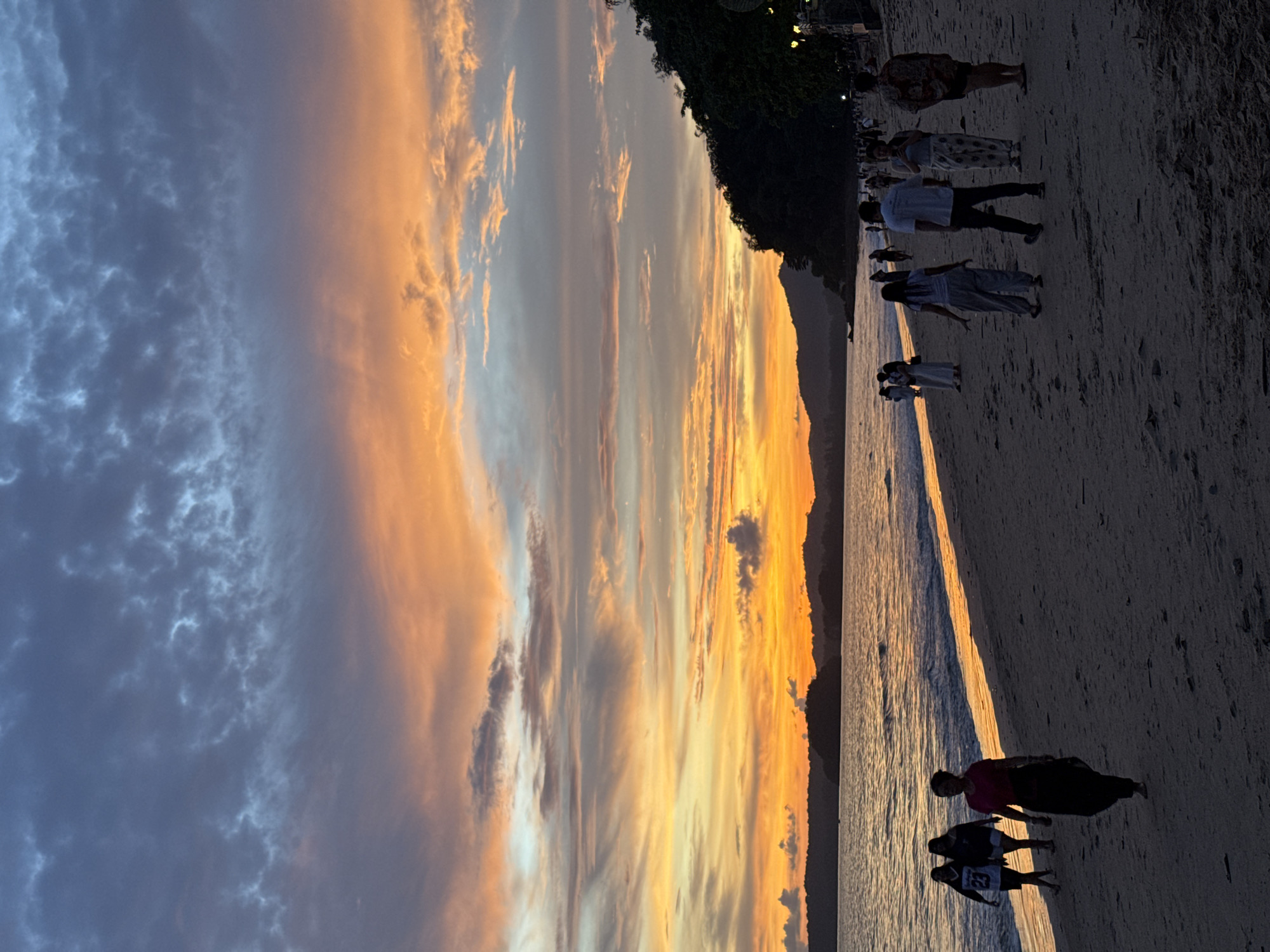 The image depicts a sunset scene on a beach in Ban Klong Haeng, Thailand. The sky is filled with dramatic clouds painted in hues of orange, yellow, and blue. A body of water, likely the sea, stretches across the lower left portion of the image, with gentle waves lapping at the shore. The beach itself is sandy, with a shoreline that curves towards the right.
Several people are visible walking along the beach, silhouetted against the colorful sky and the water. In the foreground, a woman in a pink top and dark skirt is walking towards the viewer. To her left, two younger women, also silhouetted, are walking away from the viewer. Further down the beach, a diverse group of individuals are casually strolling or standing, their figures indistinct in the fading light.
The background features a low-lying, tree-covered hill or coastline that forms a dark silhouette against the bright sky. The overall impression is one of a serene evening at a popular beach location.