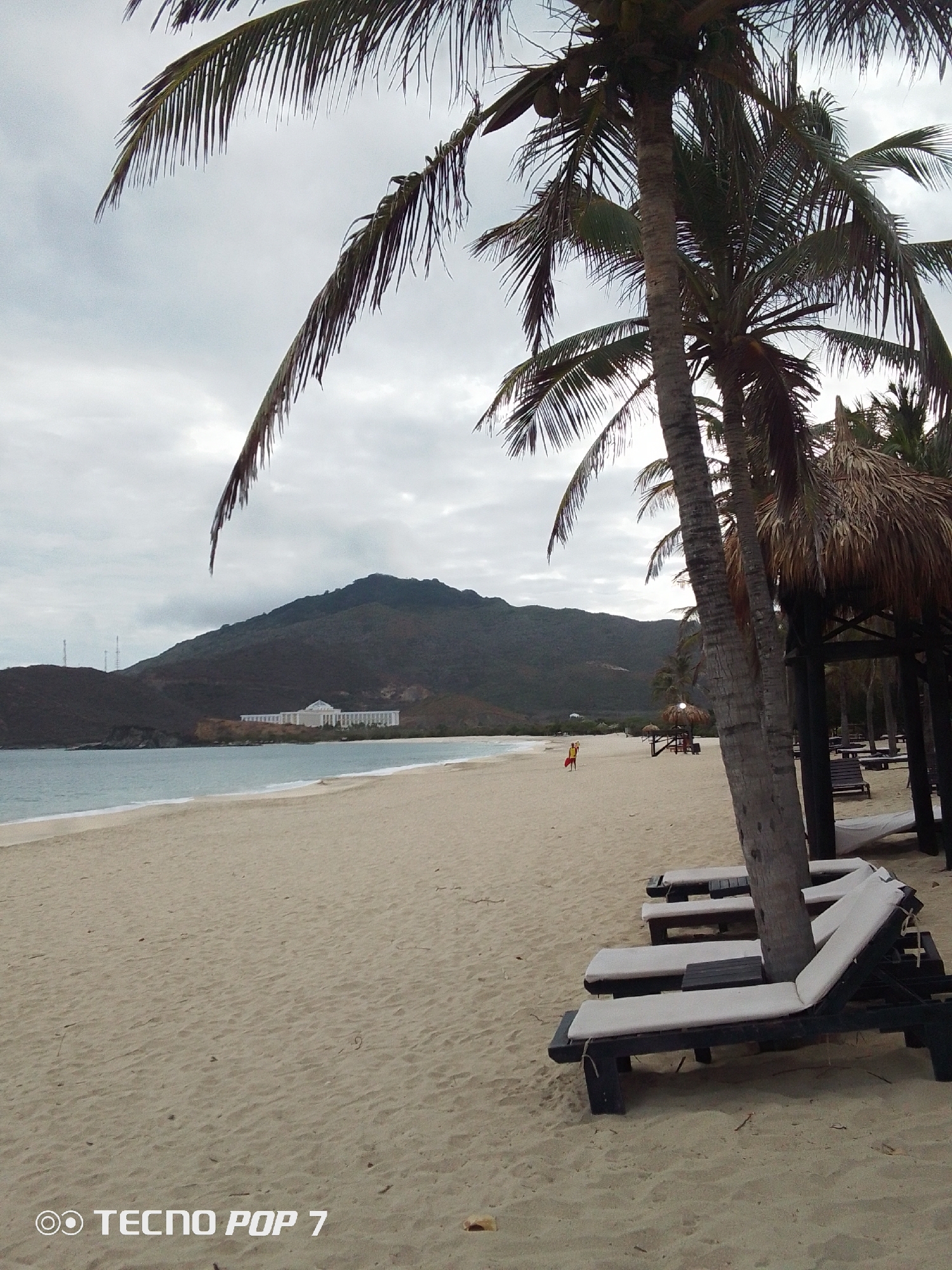 The image depicts a beach in Pedro González, Venezuela. The scene is dominated by a sandy beach, meeting the water on the left side of the frame. In the background, there's a mountain with a building near its base. The sky is overcast. Several palm trees are present, some of which obstruct the view. In the foreground, there are beach loungers. A person wearing red is walking on the beach in the mid-distance. Structures with thatched roofs are visible near the loungers. The text "TECNO POP 7" is printed at the bottom of the image.