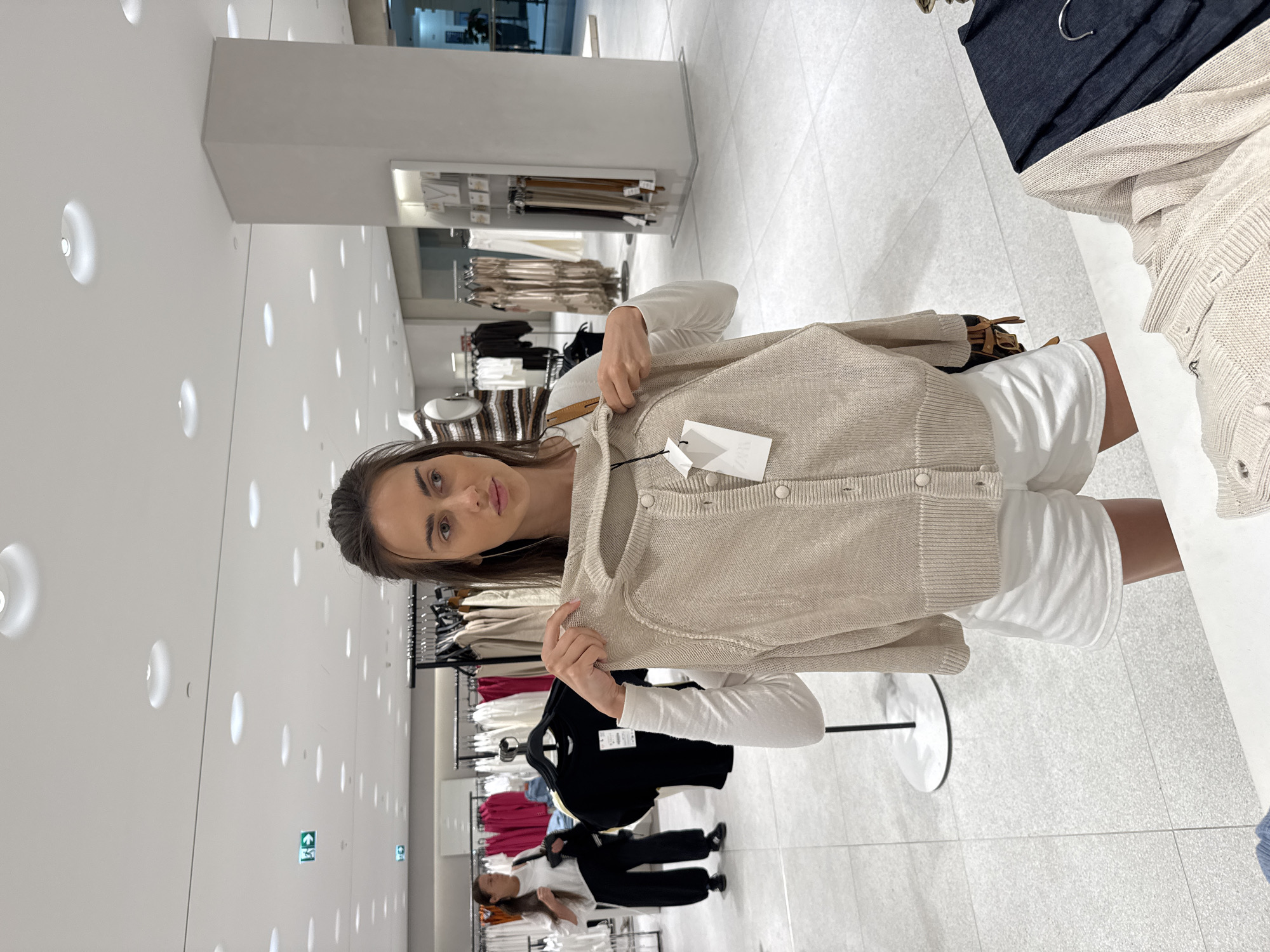 The image is taken inside a clothing store. A woman is holding up a beige cardigan with tags in the center of the frame. She has brown hair and fair skin. She is wearing a white long-sleeved shirt and white shorts. In the background, there are clothing racks with various garments in different colors. Other shoppers can be seen browsing the racks. The store has bright, white lighting with recessed lights in the ceiling. The floor is a light gray tile.