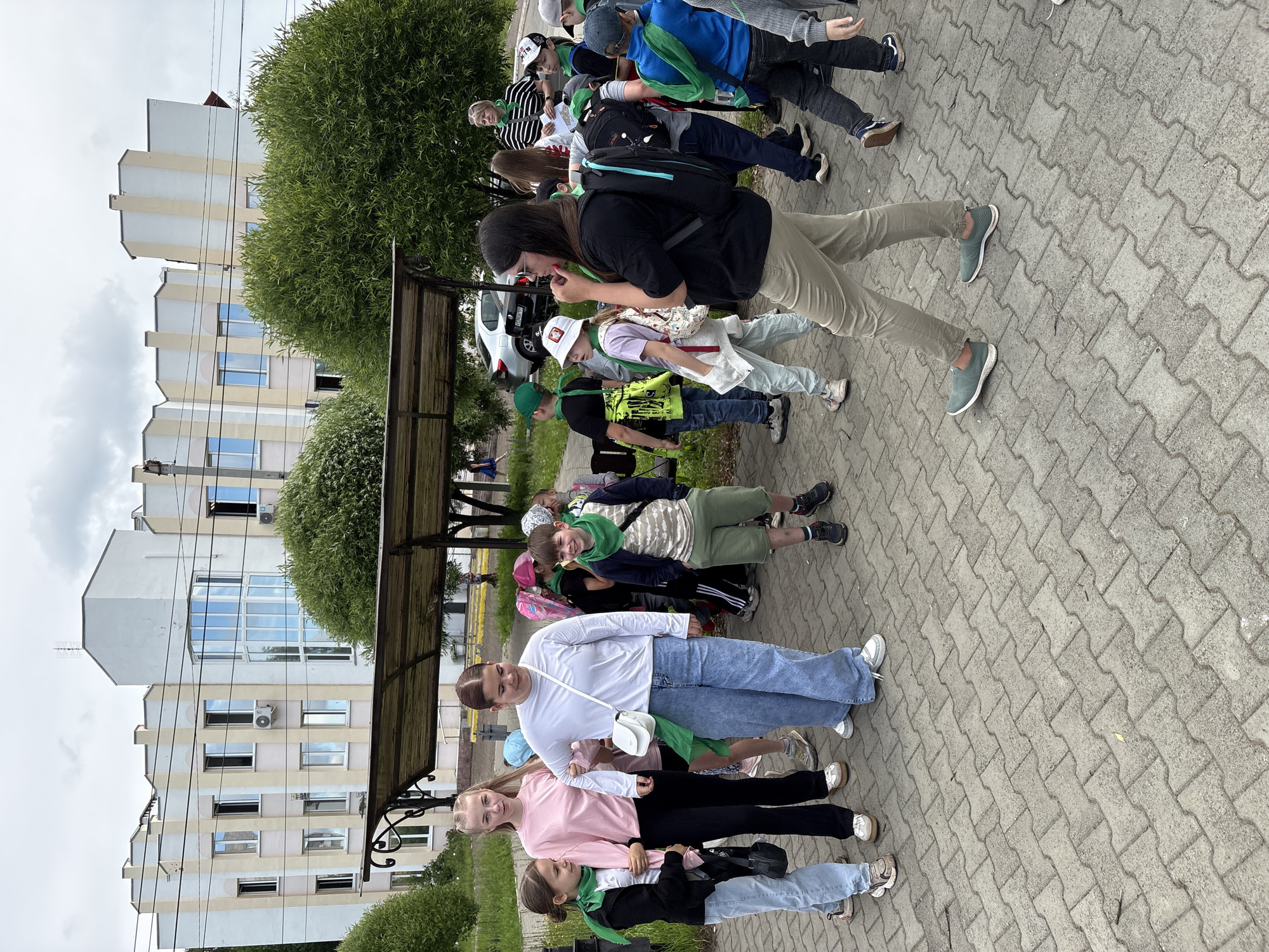 The image depicts a group of children and a few adults gathered on a paved area in front of a building. The building appears to be a multi-story structure with a light-colored facade, featuring several windows and balconies. There are also trees and bushes surrounding the area. The ground is covered with interlocking paving stones.
Several children are wearing green scarves, suggesting they might be part of a school trip or an organized activity. Some children are carrying backpacks. One adult, a woman wearing sunglasses and a black top, is standing and looking towards the right, with her hand near her chin. She is wearing khaki pants and green shoes. Another woman in a striped shirt is visible in the background, facing the children.
The context of the image being in Chaikovsky, Russia, is not directly discernible from the visual elements alone. However, if this location is provided as fact, it implies the scene is set in a public space within that Russian city. The overall atmosphere suggests an outdoor gathering, possibly a break during a field trip or an educational excursion.
