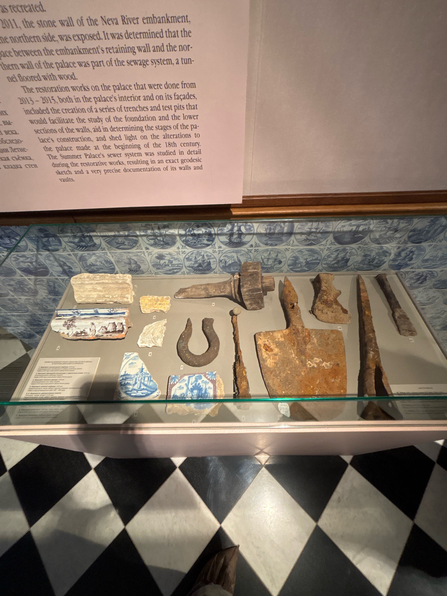 The image shows a display case in a museum containing historical artifacts. The artifacts are laid out on a light-colored surface and include fragments of pottery with blue and white decorative patterns, a rusted horseshoe, and several metal tools that appear to be aged and corroded, such as a large cleaver-like tool, a hammer-like tool, and pointed metal rods. Behind the artifacts, the wall is covered with blue and white tiles, each depicting a scene with sailing ships and coastal landscapes. The display case is illuminated, and the floor in the foreground has a black and white checkered diamond pattern. The text on the wall to the left of the display case mentions restoration work on the "Summer Palace's sewer system" and its connection to the "Neva River embankment" in the city of Saint Petersburg, Russia, suggesting this display is likely located at the State Hermitage Museum or a related historical site.