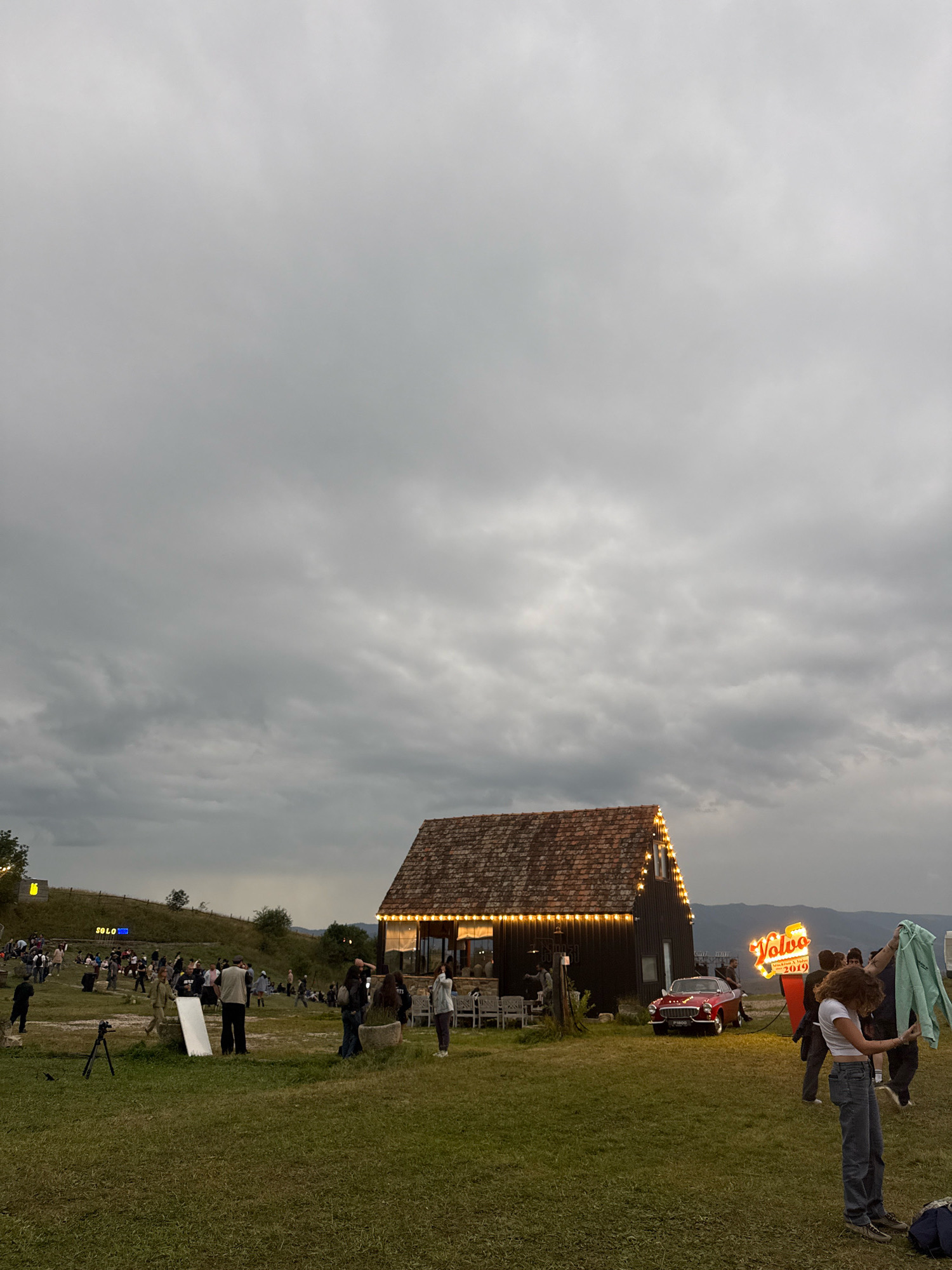 The image captures an outdoor event at Orbeti, Georgia, set against a cloudy sky. In the foreground, a grassy field is populated by several individuals engaged in various activities. To the right, a woman in jeans and a white top is holding a light blue hooded jacket. Behind her, another woman in a white t-shirt and jeans is visible.
A dark, wooden building with a tiled roof, adorned with numerous warm-toned lights, is a central feature. In front of this building, a vintage red car is parked. A prominent neon sign that reads "Volvo" in a stylized font, accompanied by text below, stands to the right of the car.
Further into the background, a crowd of people is gathered, and a hillside with a "SOLO" sign is visible. The overall scene suggests a festive or public gathering in a rural or semi-rural setting. The lighting indicates it is likely dusk or early evening.