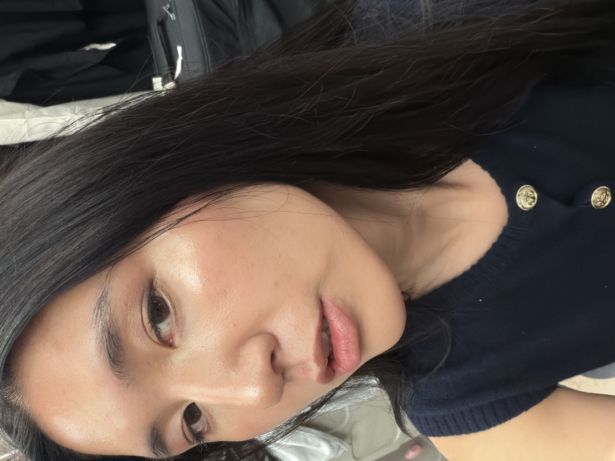The image is a close-up portrait of a young woman, taken from an upward angle, with her head tilted to the side. She has long, dark, straight hair that frames her face and extends across the left side of the frame. Her skin appears luminous and dewy, with subtle highlighting on her cheekbones. She has dark eyes, looking directly at the camera, and her eyebrows are well-defined. Her lips are slightly parted, showing a natural, soft pinkish hue. She is wearing a dark blue, possibly knitted, top with prominent gold, ornate buttons visible on the right side of the frame. The background is soft and out of focus, consisting of dark and light muted tones, suggesting an indoor setting. There are no specific architectural or outdoor elements visible to denote the location, but the context provided indicates this scene is in Paris, France. Her expression is calm and direct, possibly a slight hint of a smile. No text is visible in the image. The time of day and weather cannot be determined from the indoor setting.