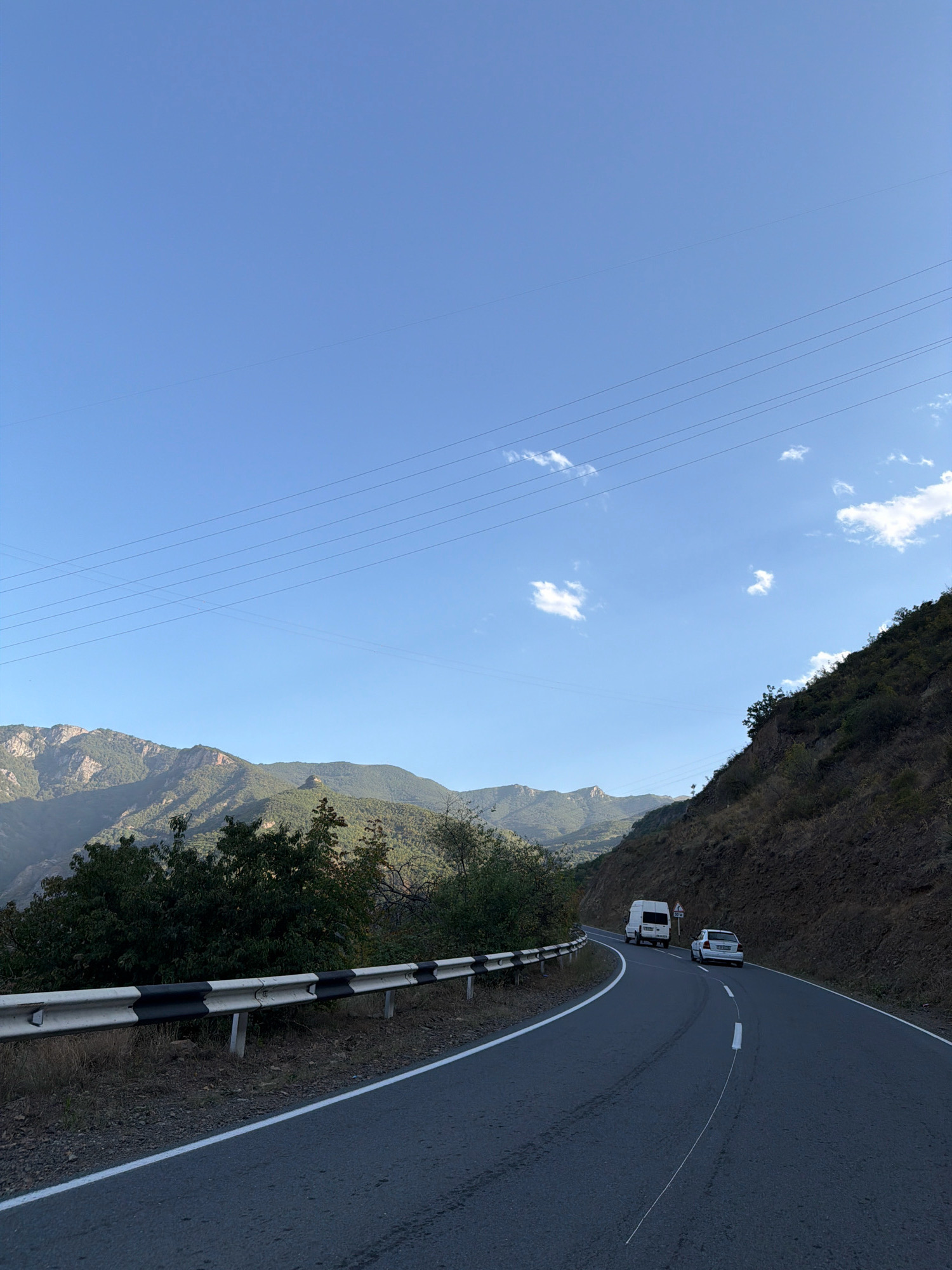 The image depicts a scenic mountain road in Tatev, Armenia. A winding asphalt road with white and black striped guardrails runs through the foreground. The road curves to the right, leading uphill and away from the viewer. On the road, a white van is driving ahead of a white car. The surrounding environment is characterized by steep, rugged mountains covered in dry vegetation and scattered trees. The sky is a clear blue with a few scattered white clouds. Overhead, power lines stretch across the frame. The overall impression is one of a remote and beautiful natural landscape.