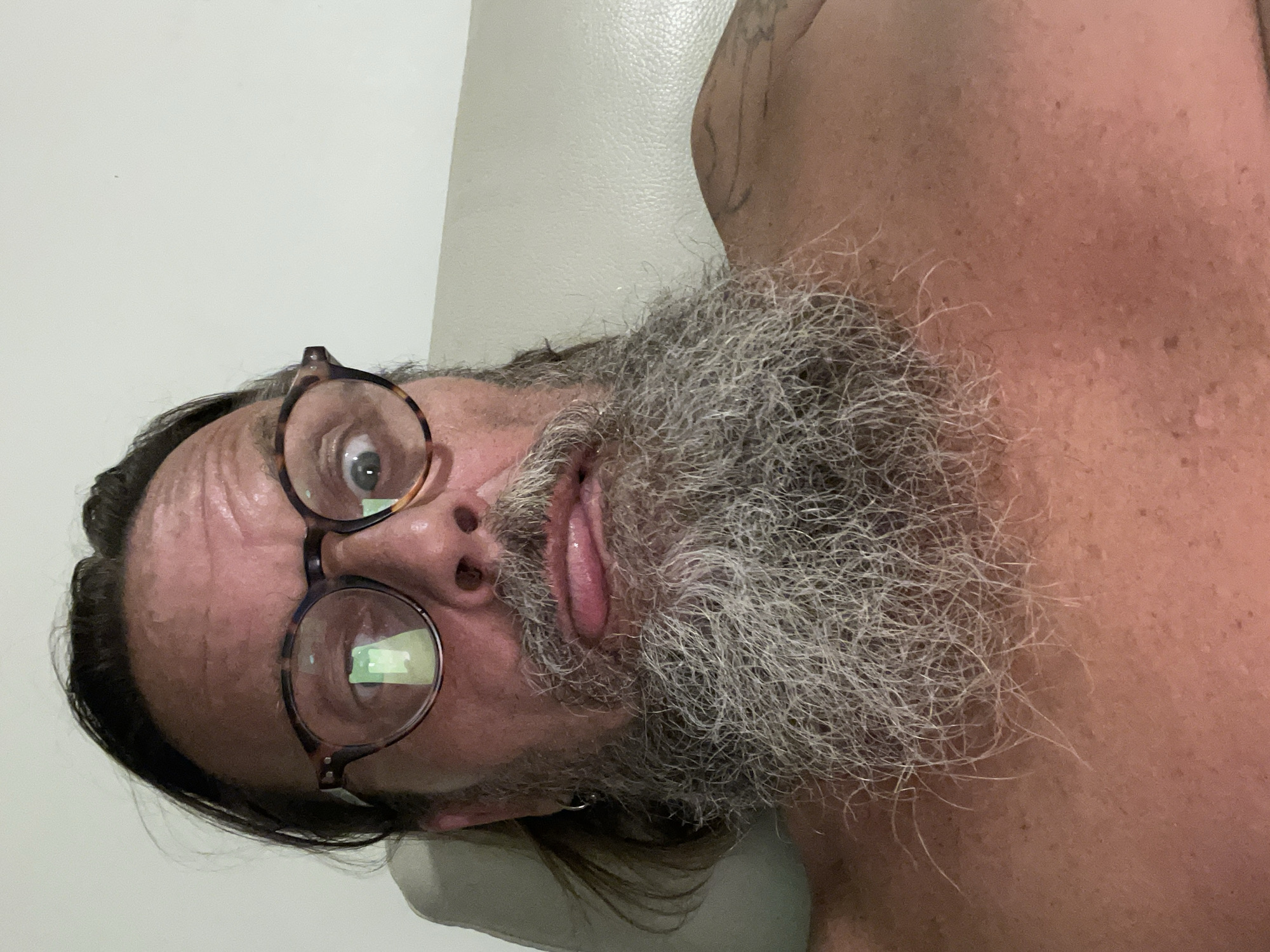 The image is a close-up selfie of a middle-aged man with a long, grey beard, wearing eyeglasses with tortoiseshell frames. He is making a funny face, sticking his tongue out slightly and widening his eyes. His hair is dark and pulled back. He appears to be shirtless. A light-colored textured surface, possibly upholstery, is visible behind him, suggesting he is indoors. There is a tattoo visible on his right shoulder. The background does not provide any specific clues to identify the city of Netanya, Israel.