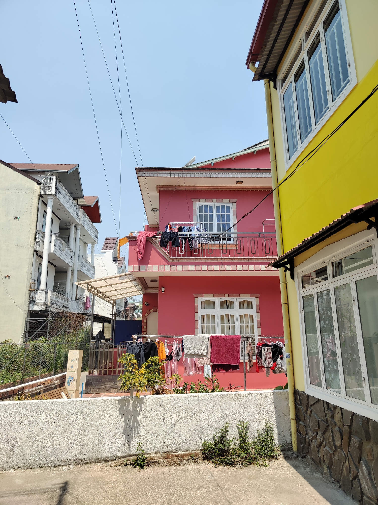 The image captures a street scene in Cam Ly - Đà Lạt, Vietnam, showcasing a residential area. A two-story pink house with white-framed windows and a balcony takes center stage, with laundry hanging on clotheslines both on the balcony and in the yard below. To the right, a yellow building with a stone facade is visible. On the left, a three-story white building with balconies can be seen. Overhead, electrical wires stretch across the clear, blue sky. A low, concrete wall runs along the front of the pink house's yard, with small plants growing near it. The street itself is concrete.