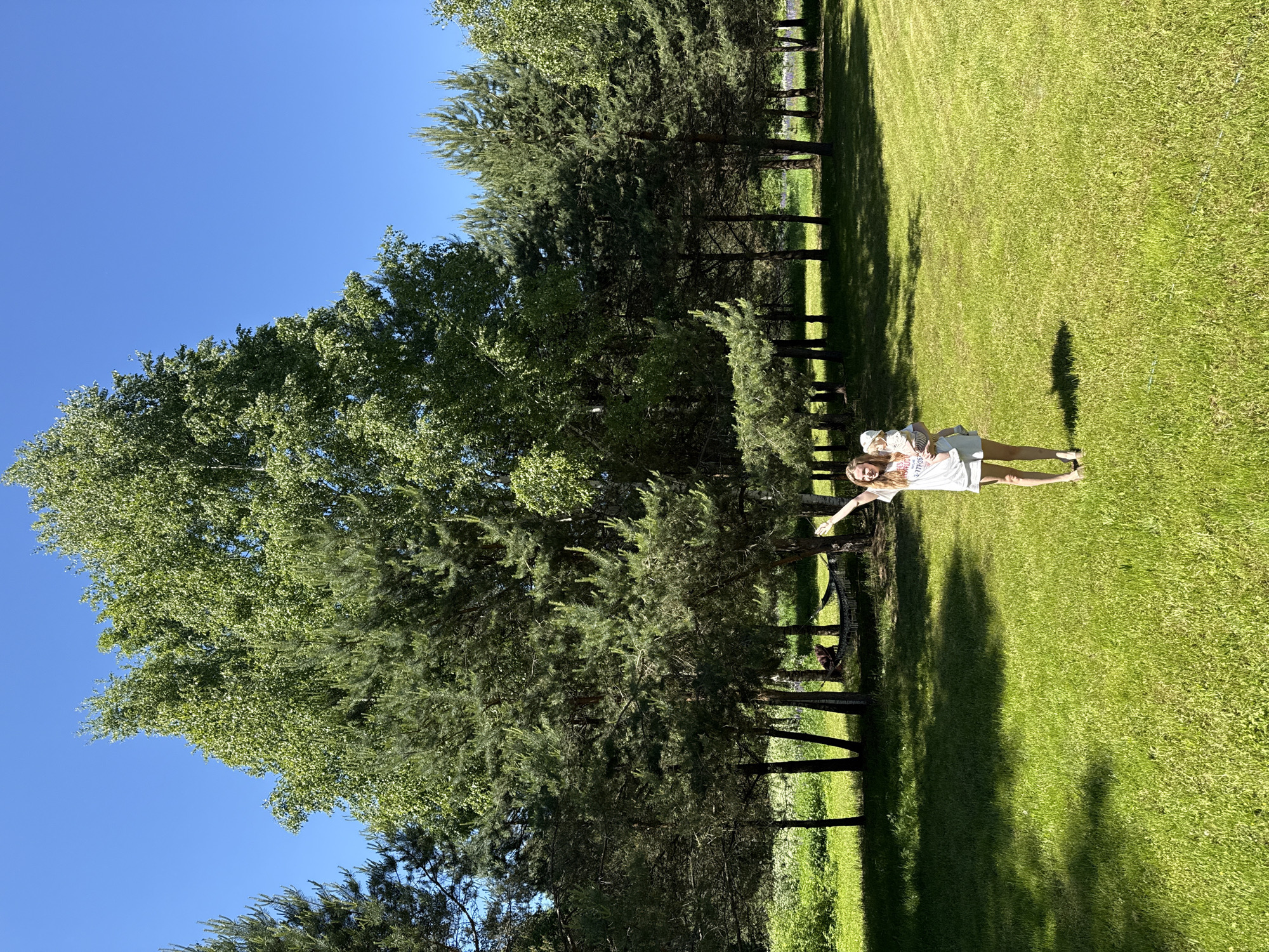 The image depicts an outdoor, sunlit scene in the location of Копнино, Russia, featuring an expansive, vibrant green grass lawn bordered by dense tree lines under a clear, cloudless blue sky.
In the foreground and slightly to the right of center, a woman stands barefoot on the grass, facing slightly left of the camera with a broad smile. She has light-colored hair and is dressed in a white short-sleeved t-shirt with an illegible dark logo or text, and light-colored shorts. Her right arm is raised and bent at the elbow, with her hand extended upward. Her left arm cradles a young child, possibly a baby or toddler, who is wearing a light-colored cap and a striped shirt. The child's head is turned slightly to the right, resting against the woman's chest.
The background consists of a dense arrangement of trees. A tall, mature deciduous tree with abundant bright green foliage, likely a birch, is centrally positioned, reaching high into the frame. Behind the woman and extending across the mid-ground is a line of coniferous trees, primarily pines, creating a thick green backdrop. Sunlight casts distinct, elongated shadows of the trees and the two individuals onto the grassy lawn, indicating a high sun position. In the mid-left background, partially obscured by tree trunks and foliage, a dark-colored hammock is visible, strung between two trees. An indistinct dark shape, possibly a person or object, is visible within or adjacent to the hammock. Faint patches of lighter green foliage and some purple flowering plants are discernible in the far background, visible through gaps in the tree line.