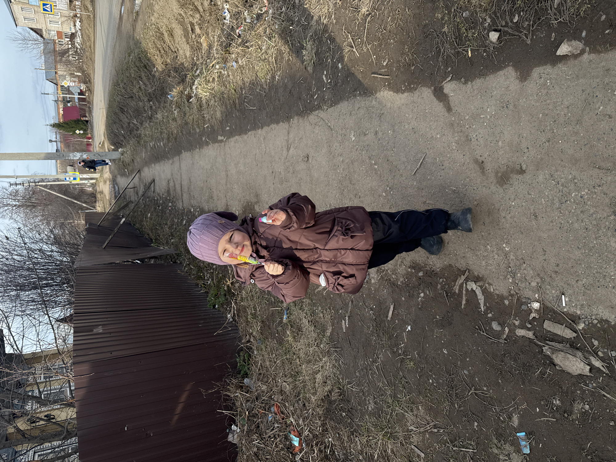 A young child in a purple hat and brown puffer jacket stands on a paved path, holding a lollipop. The path runs alongside a metal fence and dry, grassy verge littered with debris. In the background are buildings and a street scene under a partly cloudy sky. The location appears to be in a residential area. There is a crossing sign visible in the top left corner. The image suggests an urban or suburban environment.