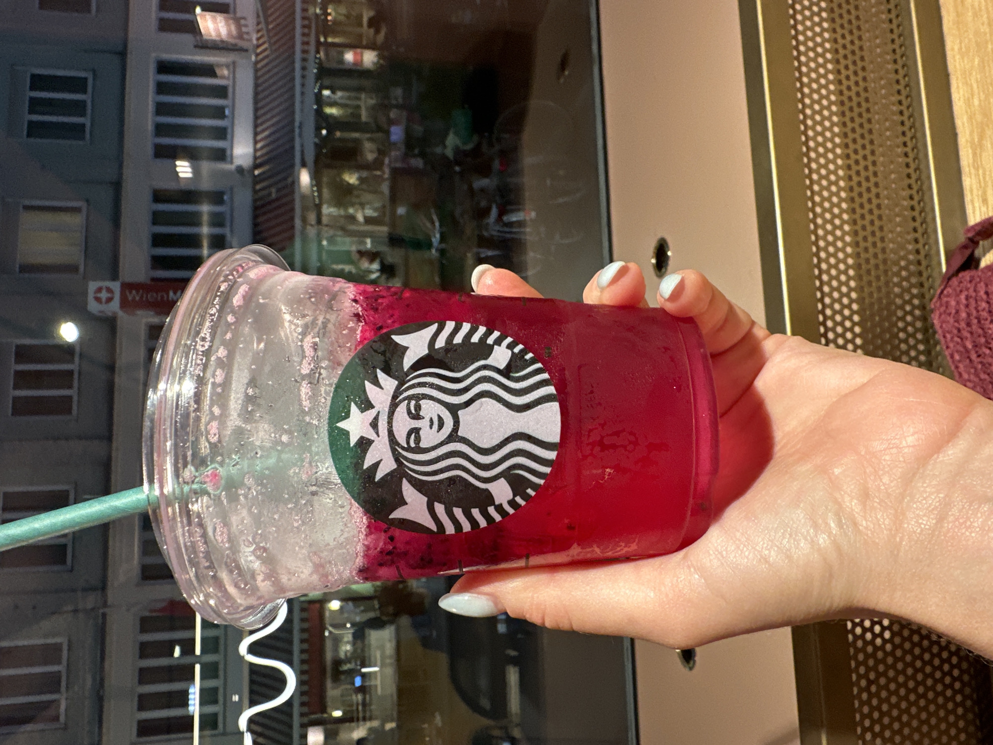The image shows a close-up of a hand holding a Starbucks cup filled with a pink, icy beverage. The Starbucks logo is visible on the cup. In the background, there is a building with windows and awnings, and a sign that reads "Wien M." This sign, in conjunction with the architecture, suggests the location is Vienna, Austria. The scene appears to be set at dusk or nighttime, as evidenced by the artificial lighting in the background.