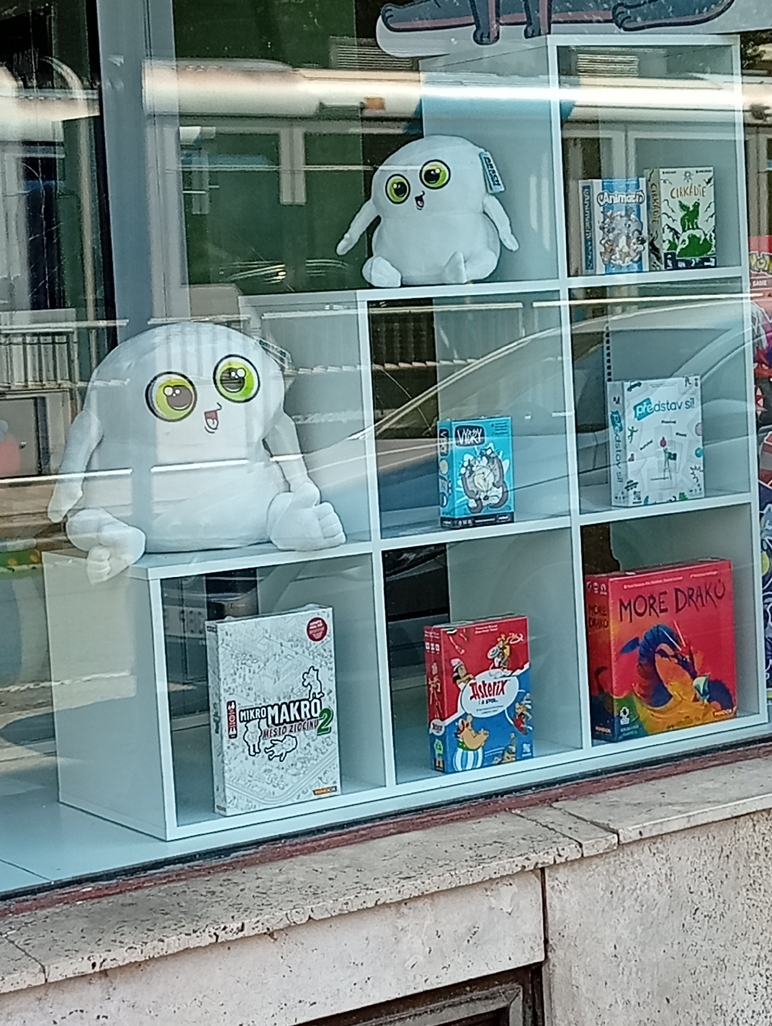 A window display shows two large plush toys and several board games. The toys are white, round, and have large green eyes with black pupils. The toy on the left is larger and appears to be sitting with its arms resting on its lap. The toy on the right is smaller and is standing. Both toys have a black tag attached to them.
The board games are displayed on white shelves. From left to right, top row: "Animalia", "Circadié", and "Představ si!". Bottom row: "Mikro Makro 2 Mesto Zlocinu", "Asterix & Obelix", and "More Draku". The display is located in Brno, Czechia.