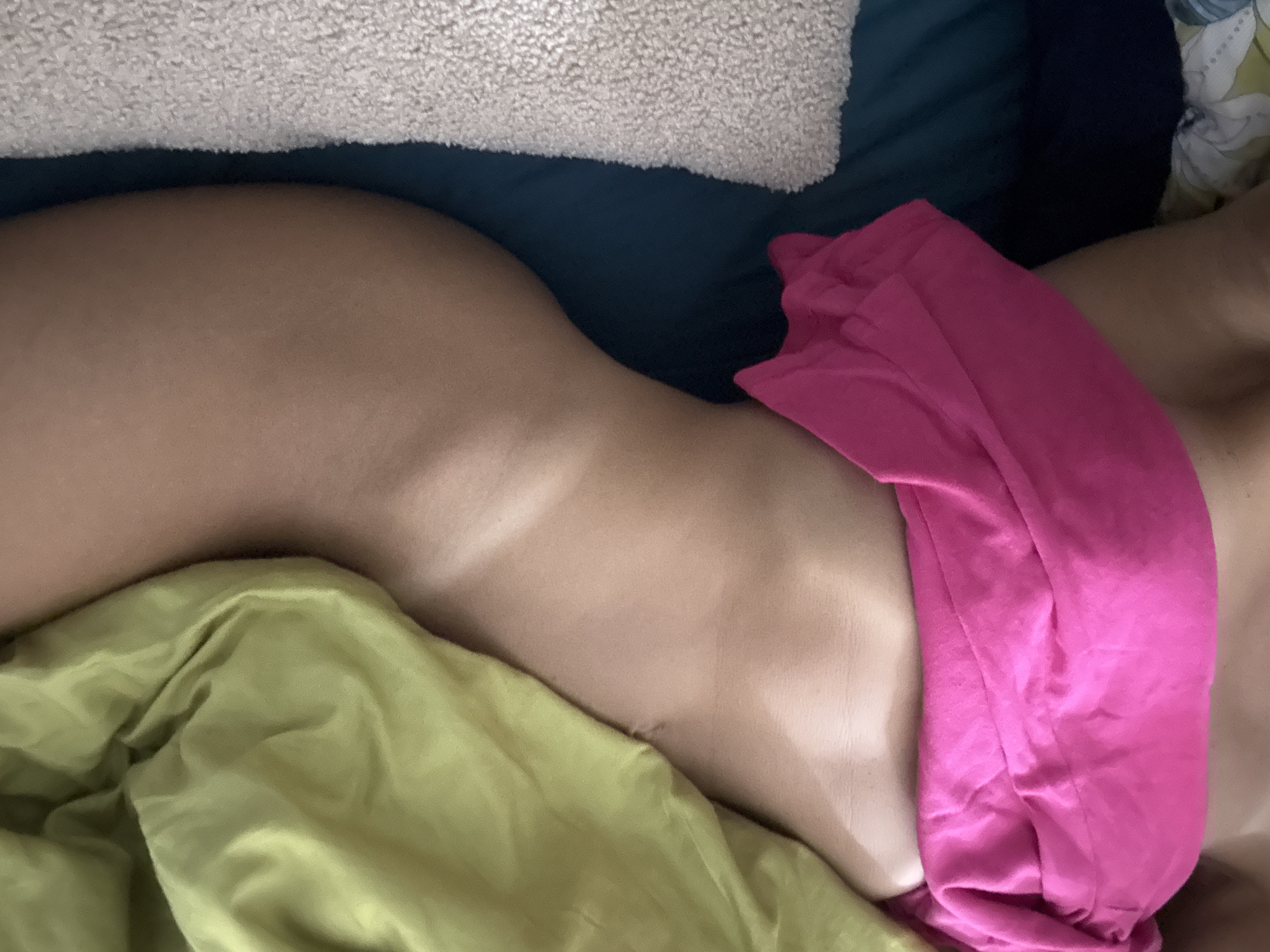 The image is a close-up, horizontal shot focusing on a person's torso and hip area, covered partially by bright pink and lime green fabric. The person is lying down, as suggested by the presence of bedding, including a textured, light-colored pillow at the top and a dark blue surface beneath it. The skin tone is light. The scene appears to be indoors, possibly in a bedroom, given the bedding. The lighting is soft, and there are no clear indicators of the time of day or weather. No text is visible in the image. The overall mood is intimate and subdued due to the close cropping and soft focus on the textures of skin and fabric.