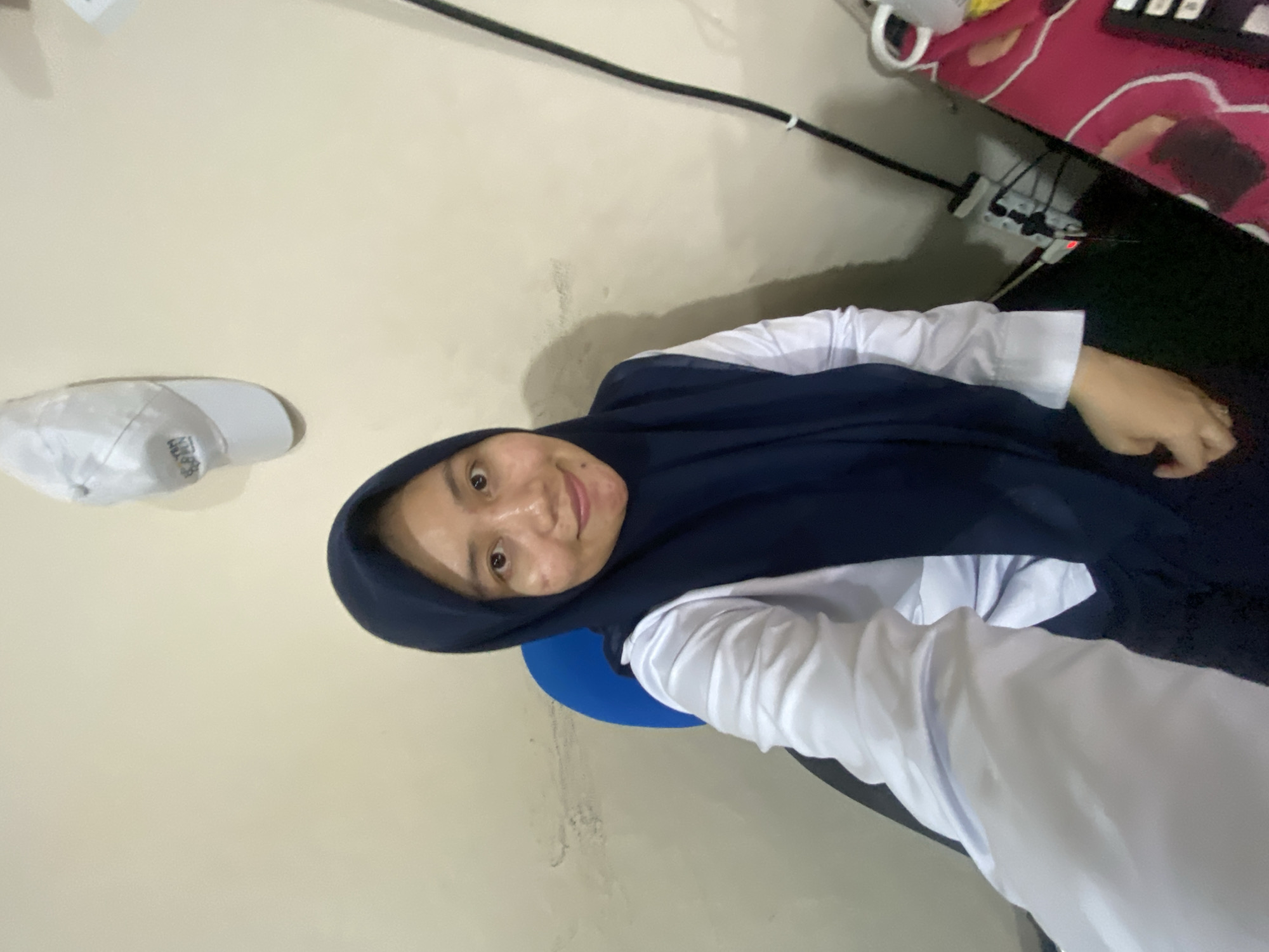 The image depicts a young woman wearing a dark blue hijab and a white collared shirt, seated in a blue office chair. She is looking directly at the camera with a slight smile. The background is a plain, light-colored wall with some visible discoloration and marks. A white baseball cap with lettering is affixed to the wall above and slightly to the left of her head. To the right of the woman, a black cable runs down the wall to a power strip with multiple outlets, one of which is illuminated red, indicating it is active. A portion of a patterned pink surface, possibly a desk or fabric, is visible in the lower right corner of the frame, with some electronic devices and cables on it. The lighting appears to be artificial and casts soft shadows. The overall impression is of an indoor, casual setting, likely a home office or study area, possibly in Banjarbaru, Indonesia.