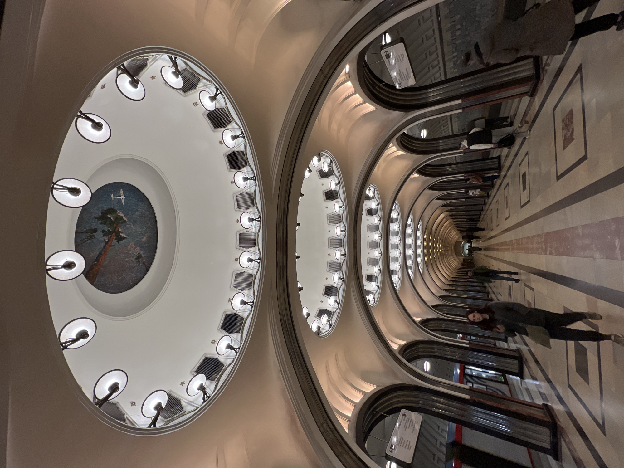 The image shows a perspective shot inside a Moscow metro station in Russia. The ceiling is adorned with repeated arched designs, each housing a row of lights. At the end of the tunnel is a central round structure on the ceiling featuring a mural. Several people can be seen walking through the metro station. The station is well-lit.