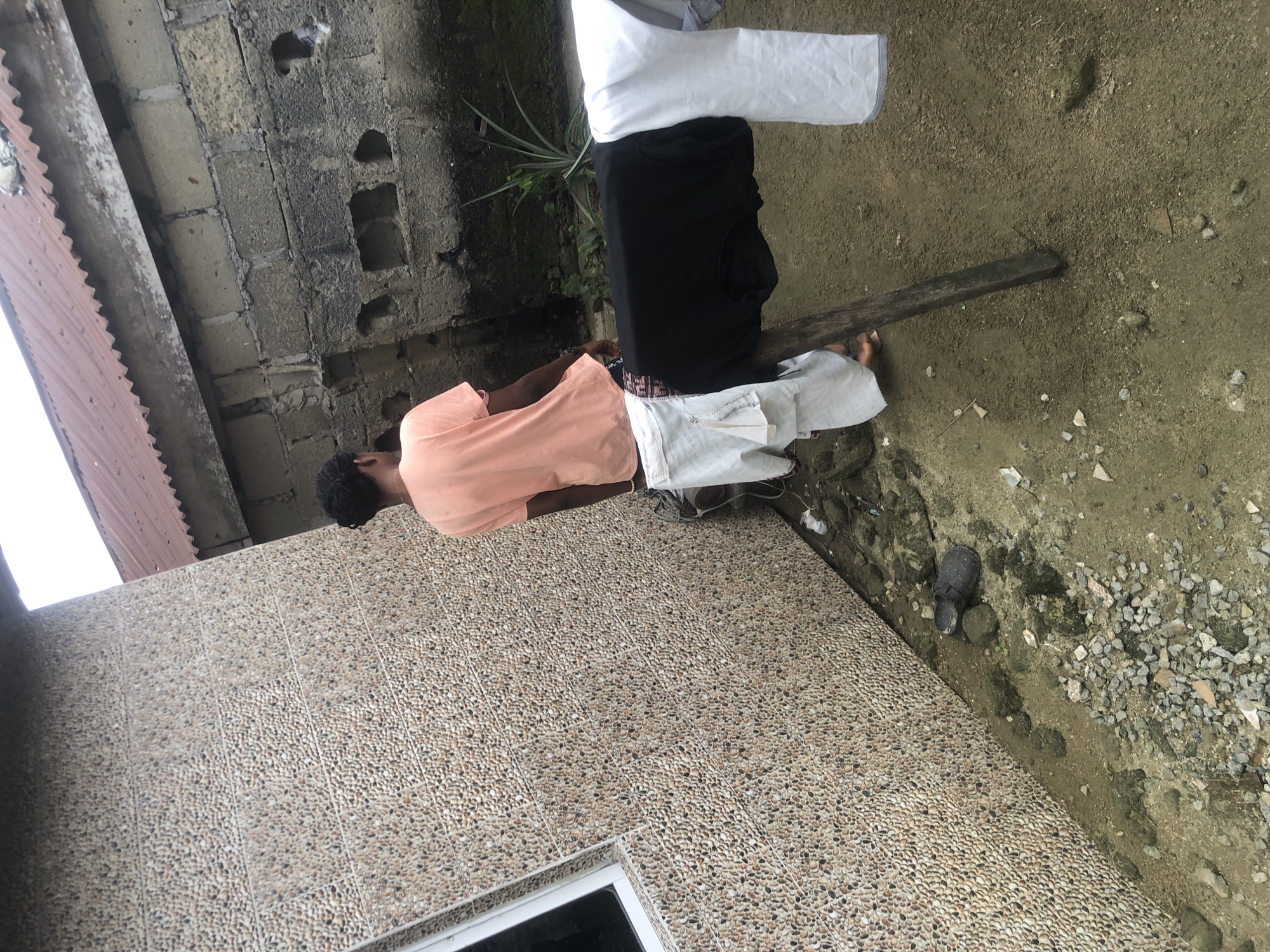 A young Black male, wearing a peach-colored t-shirt and patterned shorts, stands next to a pebble-textured wall in Ado, Nigeria. He is facing away from the camera, partially obscured by a clothesline strung between two wooden poles. On the clothesline hang a black t-shirt and two white garments, likely clothing items. The ground is dirt and strewn with small stones, gravel, and some scattered debris, including a single dark-colored shoe near the wall. In the background, a dilapidated brick wall with visible holes and a corrugated metal roof suggest an outdoor, possibly undeveloped, area. A small, spiky green plant grows near the brick wall. The overall environment appears to be a simple, outdoor domestic setting.