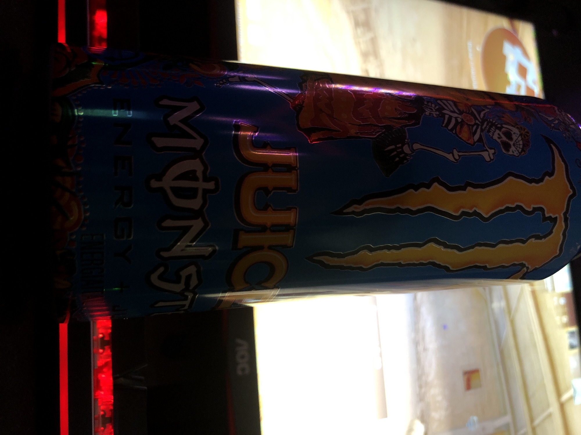 This close-up image, captured in the city of Beryozovka, Russia, displays a Monster Energy "Juice Monster" can standing upright in the foreground, positioned slightly left of center. The can features a blue base, an orange and yellow Monster logo, and an illustration of a skeleton holding a scythe. Strong reflections, displaying purple and orange hues, are visible on the can's surface, particularly on the left side and over the "JUICE" text. The can rests on a dark, flat surface. Directly in front of and to the right of the can's base, a strip of red LED lighting is visible. In the blurred background, a brightly illuminated monitor screen displays what appears to be a video game or animated content. The left side of the screen shows a partial orange circular logo with white text on a light brown background, while the right side reveals a brightly lit, empty indoor environment with light-colored walls and ceiling, featuring a small, blurry orange square object on a far wall. The lower bezel of the monitor is visible, displaying a white "AOC" manufacturer logo on the right side. The overall scene is dimly lit, with the monitor screen serving as the primary light source.