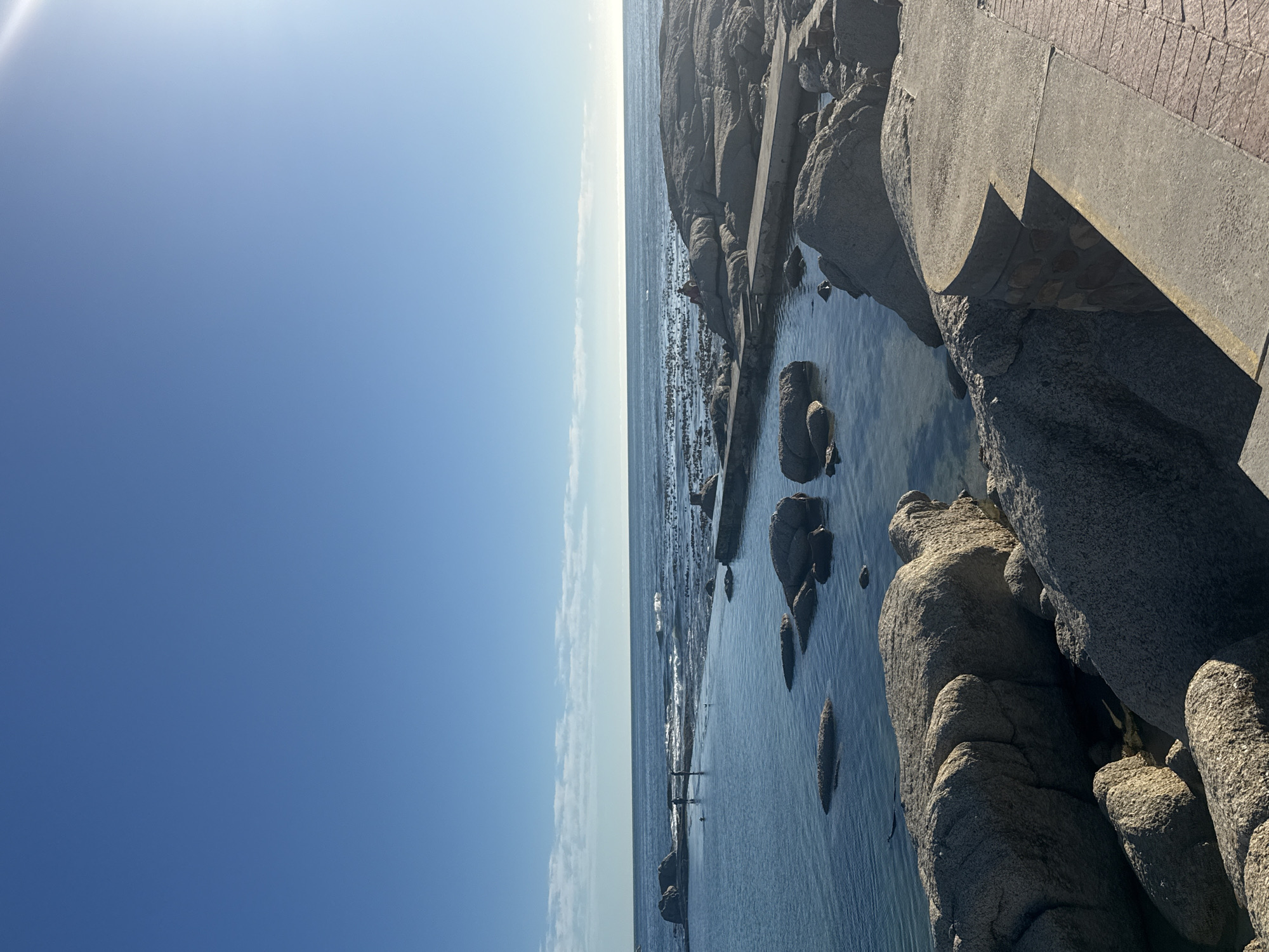 The image depicts a coastal scene in Cape Town, South Africa. The sky is predominantly blue with some visible sun glare. The ocean meets the sky with a narrow band of lighter-colored water. The foreground features large gray rocks, some of which are partially submerged in the water. A concrete structure or walkway is also visible, bordering the rocks. There are figures standing in the shallow water.