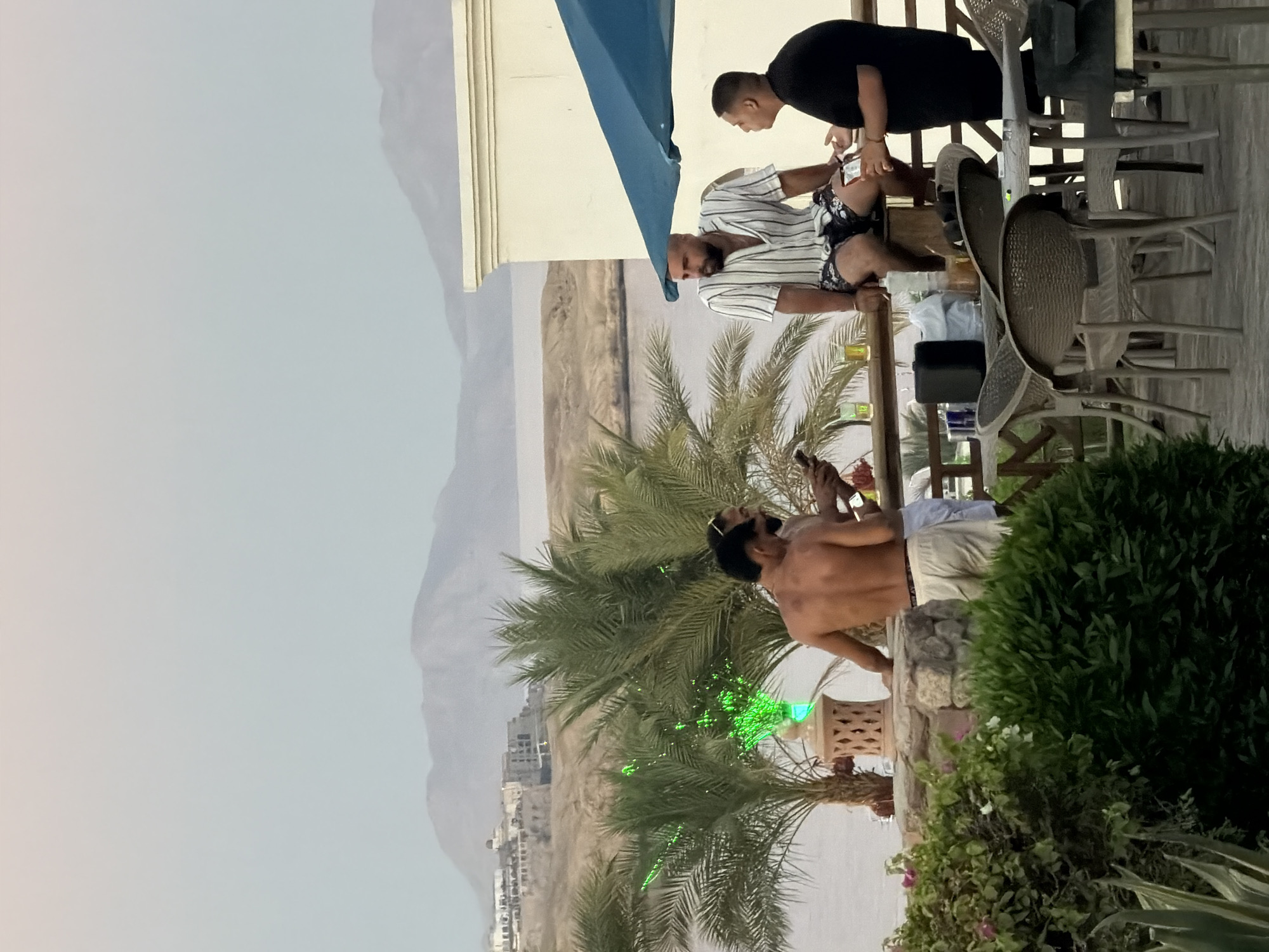 The image depicts an outdoor scene, likely a resort or restaurant patio, in Sharm El Sheikh, Egypt. The background features arid, mountainous terrain and a body of water, possibly the Red Sea, with a settlement visible on the coastline. In the foreground, several men are present. Two men are standing near a decorative stone structure with a bright green light, one with his back to the camera, showing his torso. Another man is seated on a railing, wearing a striped short-sleeved shirt and patterned shorts. He is engaged in conversation with a man standing next to him, who is wearing a black t-shirt. There are palm trees and lush green foliage framing the scene. Tables and chairs are set up on the patio, with drinks visible on a table. The sky is overcast with a gradient of pale pink to light blue.