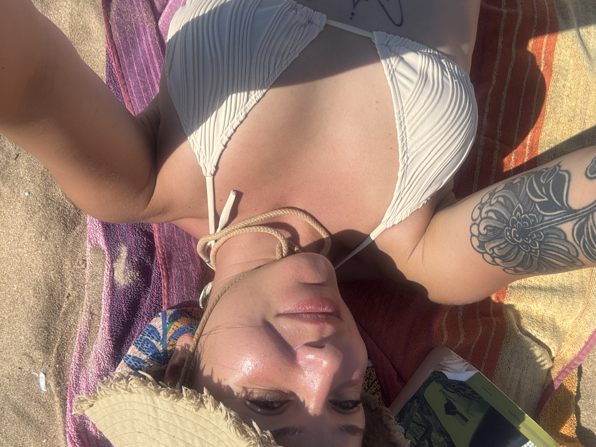 A woman is lying on a colorful beach towel on the sand. She is wearing a white bikini and a straw hat. The woman has a tattoo on her left leg. A book is also visible on the towel. The scene appears to be a sunny day at a beach, likely in Sozopol, Bulgaria, given the context. The woman is looking up at the camera.